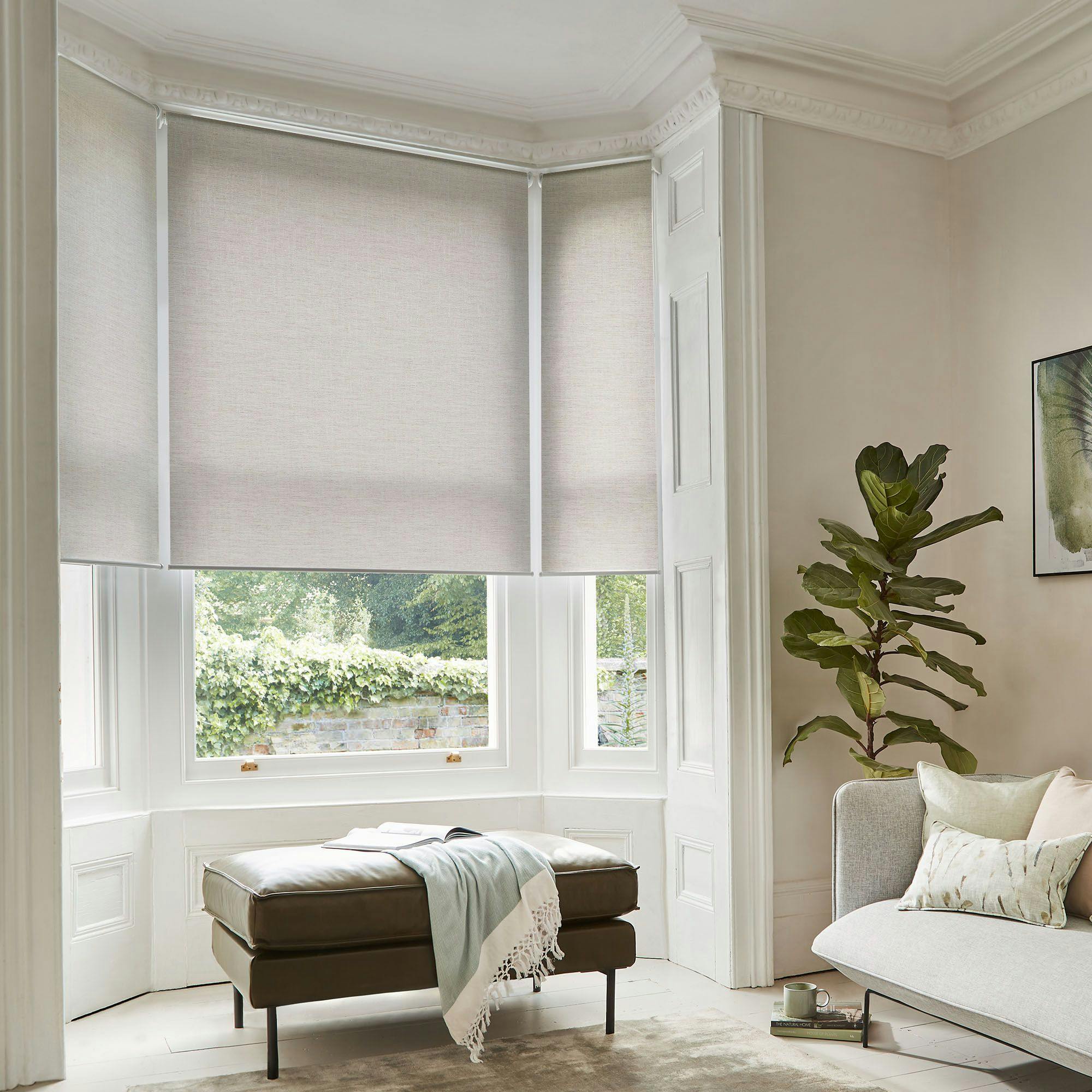 A cushioned bench draped with a blanket rests by a large bay window with beige blinds, adjacent to a corner with a potted plant, in a serene, elegant room.