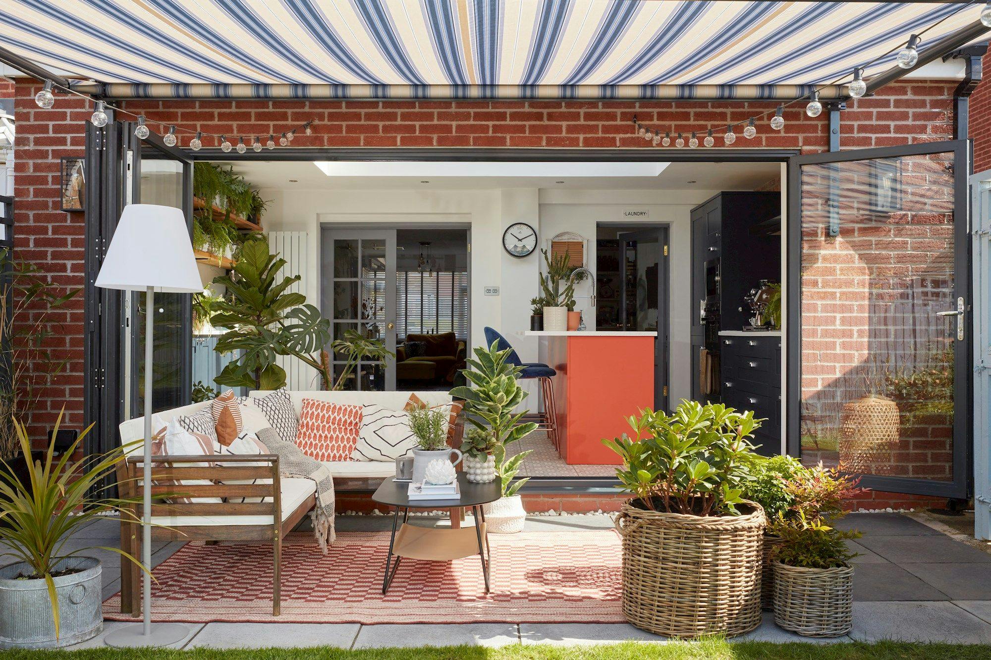 Outdoor seating area features a cozy arrangement of wooden furniture, cushions, and plants, under a striped awning. French doors lead to a modern indoor space with a kitchen.