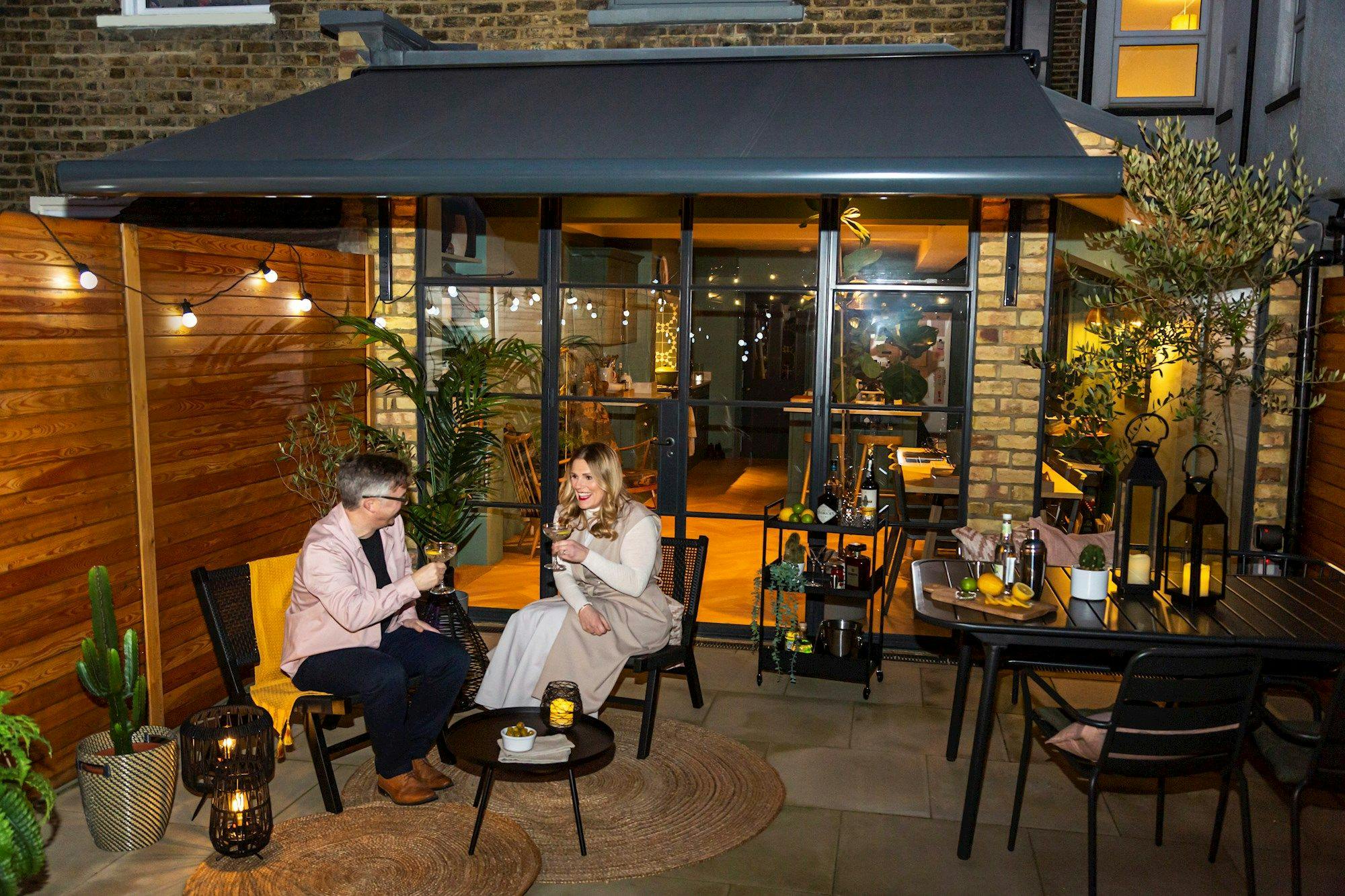 Two people sit drinking on a patio. The area is decorated with plants, string lights, and cozy furnishings. The interior, visible through glass doors, is warmly lit.