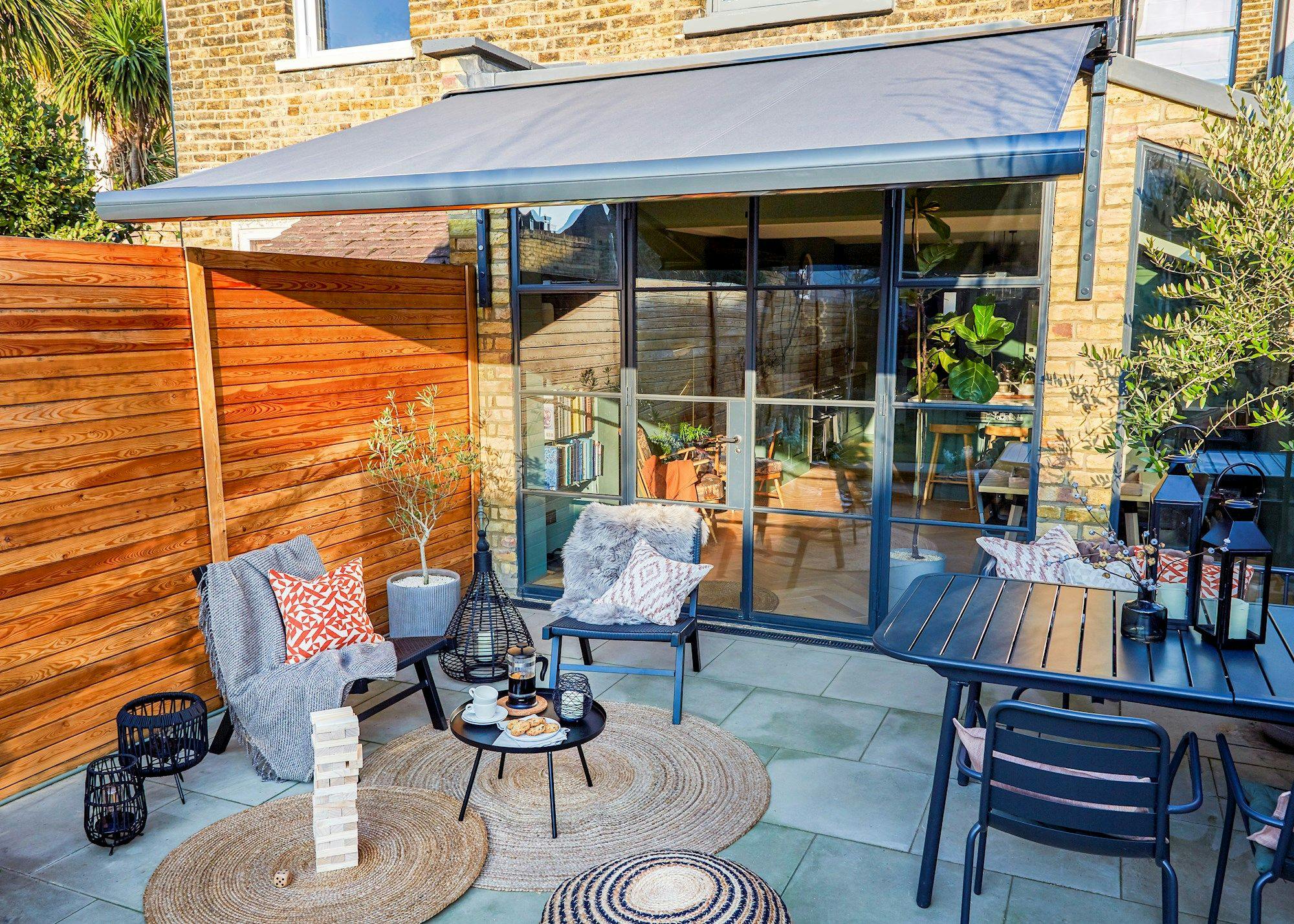 Patio with two chairs and cushions around a small table holding coffee and pastries, set near a wooden fence and glass doors leading inside a house. Large awning provides shade.