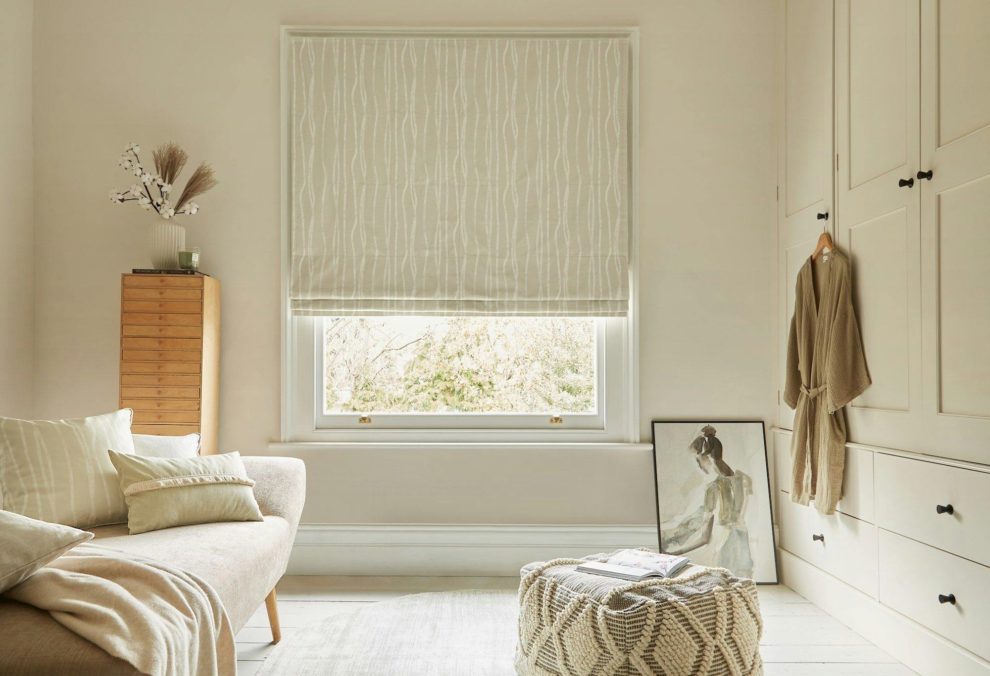 A beige sofa with cushions sits beside a window with patterned blinds, surrounded by a tall cabinet with decorative items, a pouf, a painting, and wall cabinets with a robe.