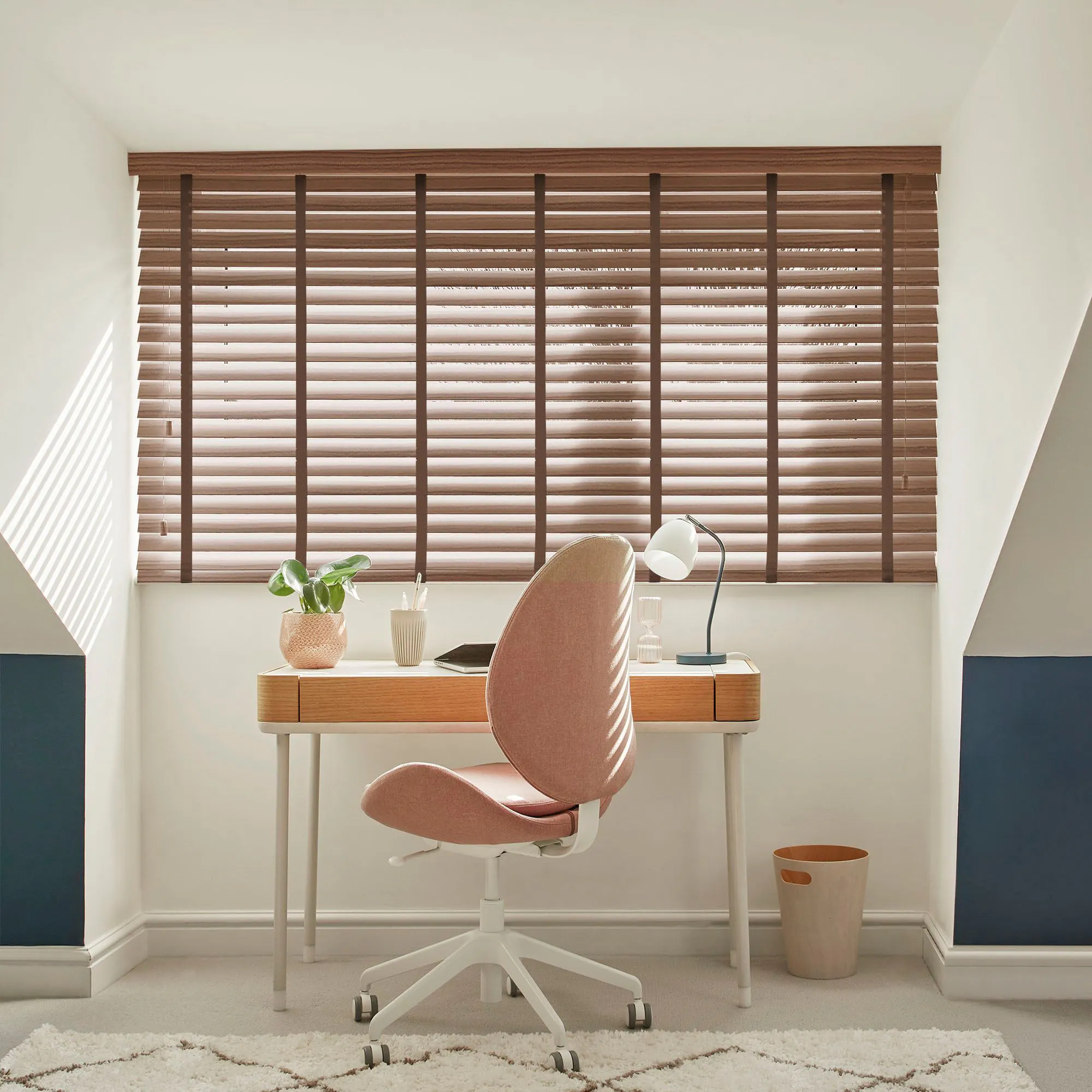 A pink office chair faces a wooden desk with a plant, lamp, and notebook. The room has wooden blinds filtering sunlight and a beige carpet, creating a cozy workspace.