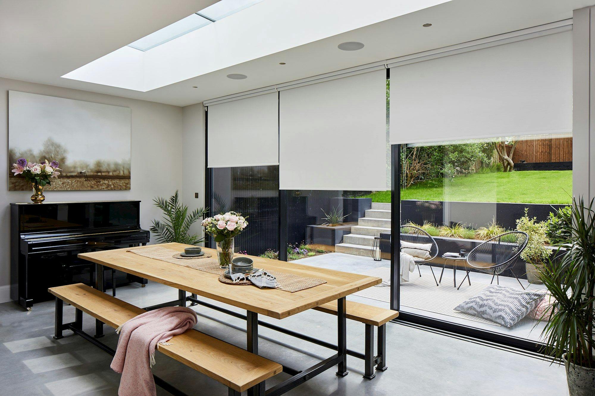 A wooden dining table with benches holds plates and flowers, facing a black piano topped with flowers. Glass doors reveal a patio with chairs, stairs, and a grassy yard.