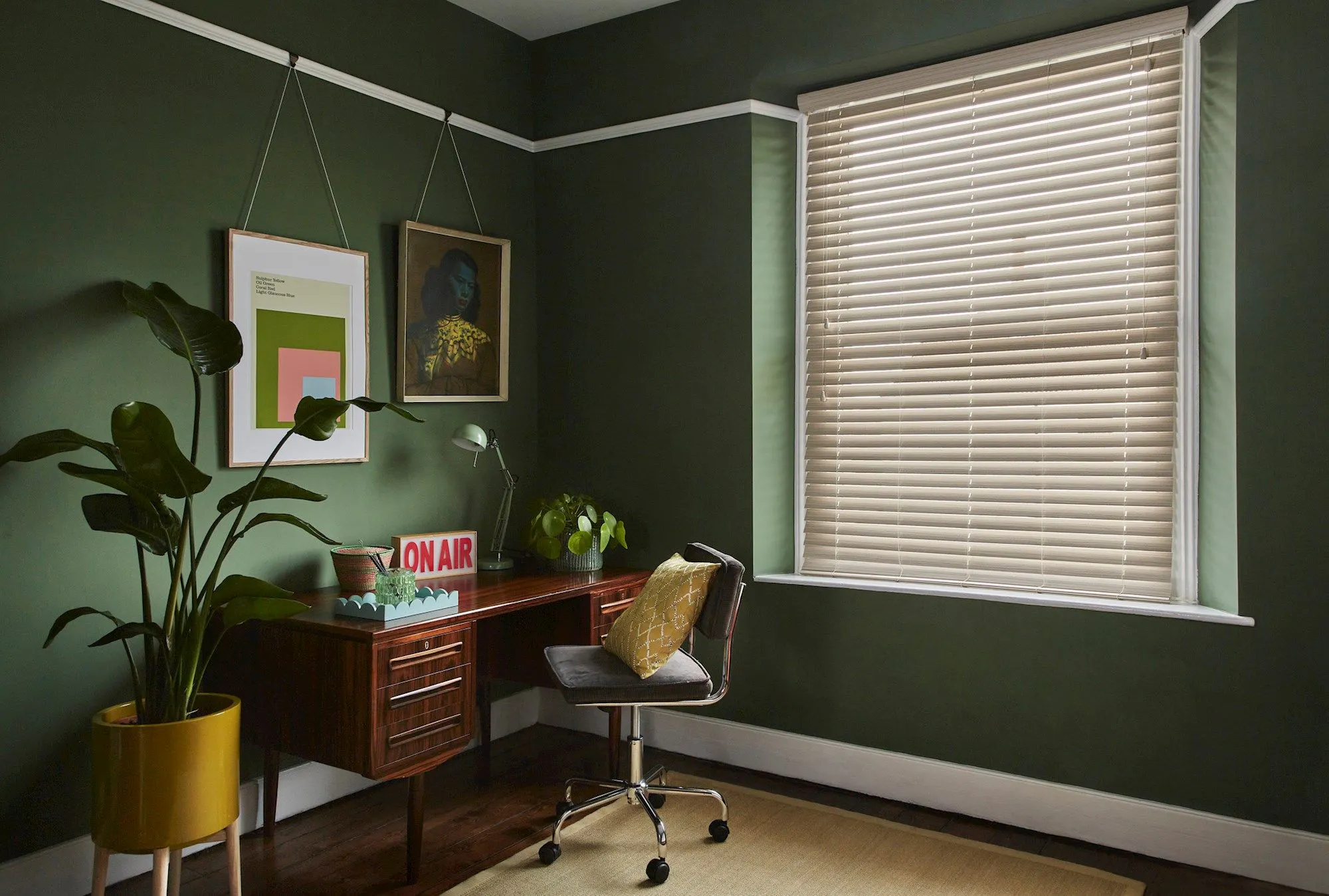 A mid-century-style dark green home office with a cream Wooden blind and a large potted plant.