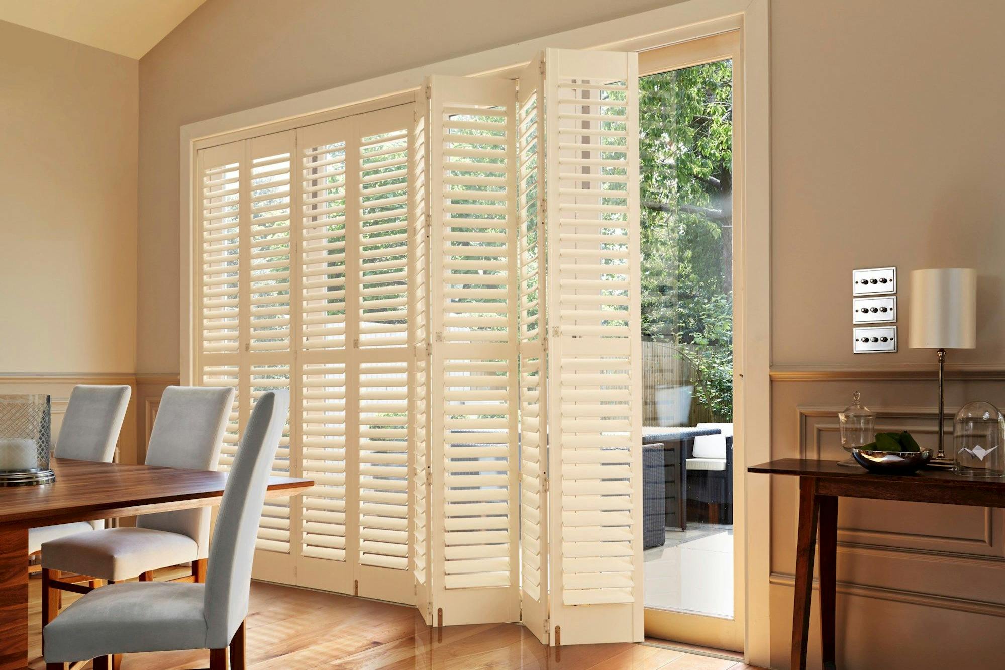 White plantation shutters partially open, revealing a garden view. Surrounding them, a dining room with a wooden table, upholstered chairs, and soft, natural light filtering through.