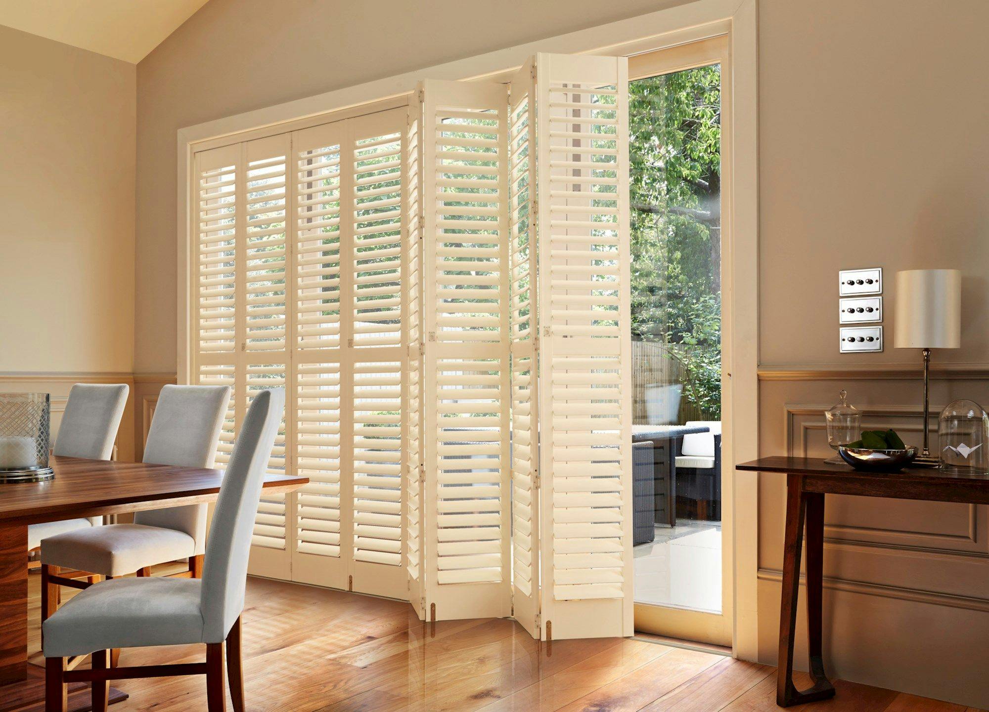 White plantation shutters partially open, revealing a garden view. Surrounding them, a dining room with a wooden table, upholstered chairs, and soft, natural light filtering through.