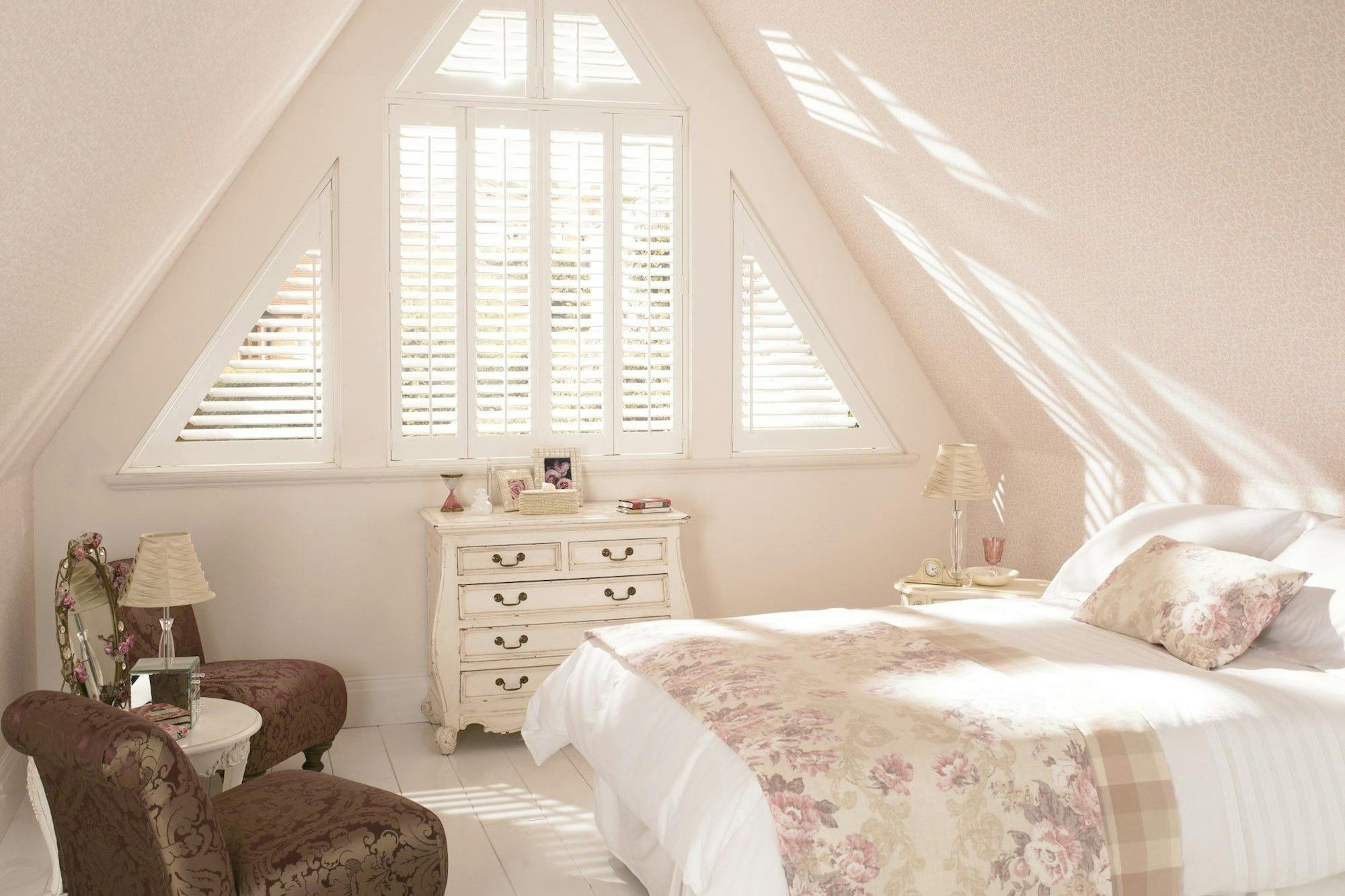 A bed with floral bedding is bathed in sunlight streaming through triangular windows with shutters, situated in an elegantly decorated attic bedroom with vintage furniture and pastel colors.