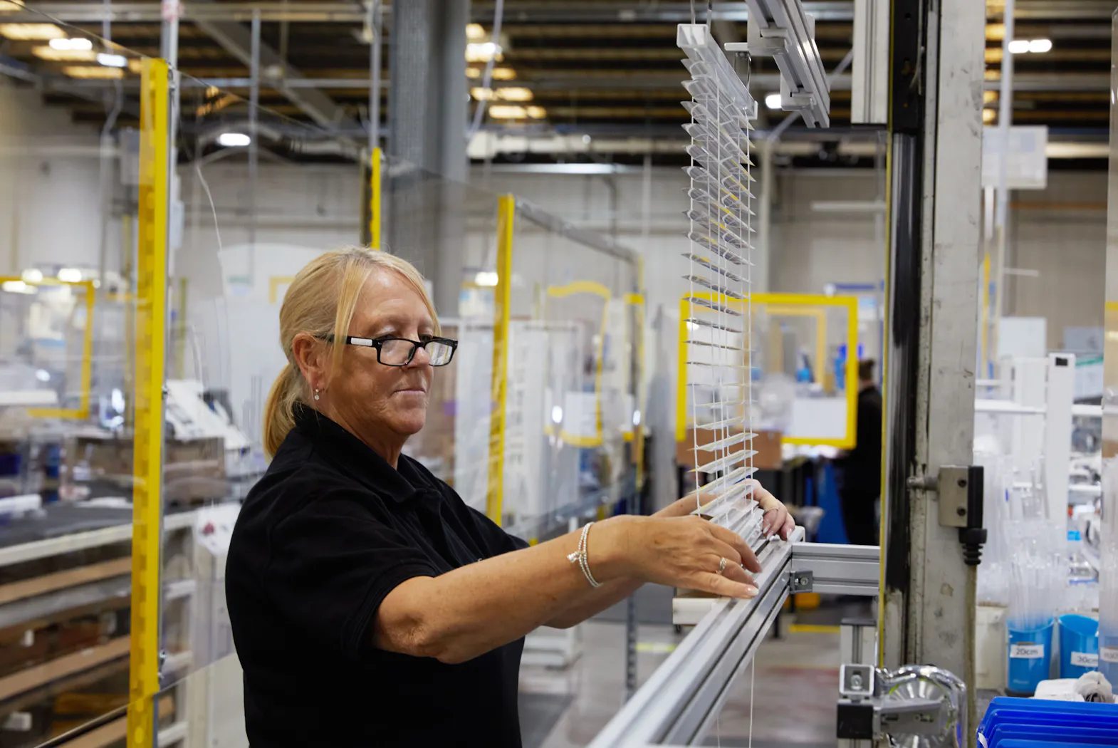 A woman adjusts window blinds in a modern factory, surrounded by production equipment and industrial shelving, with protective screens and bright overhead lighting.