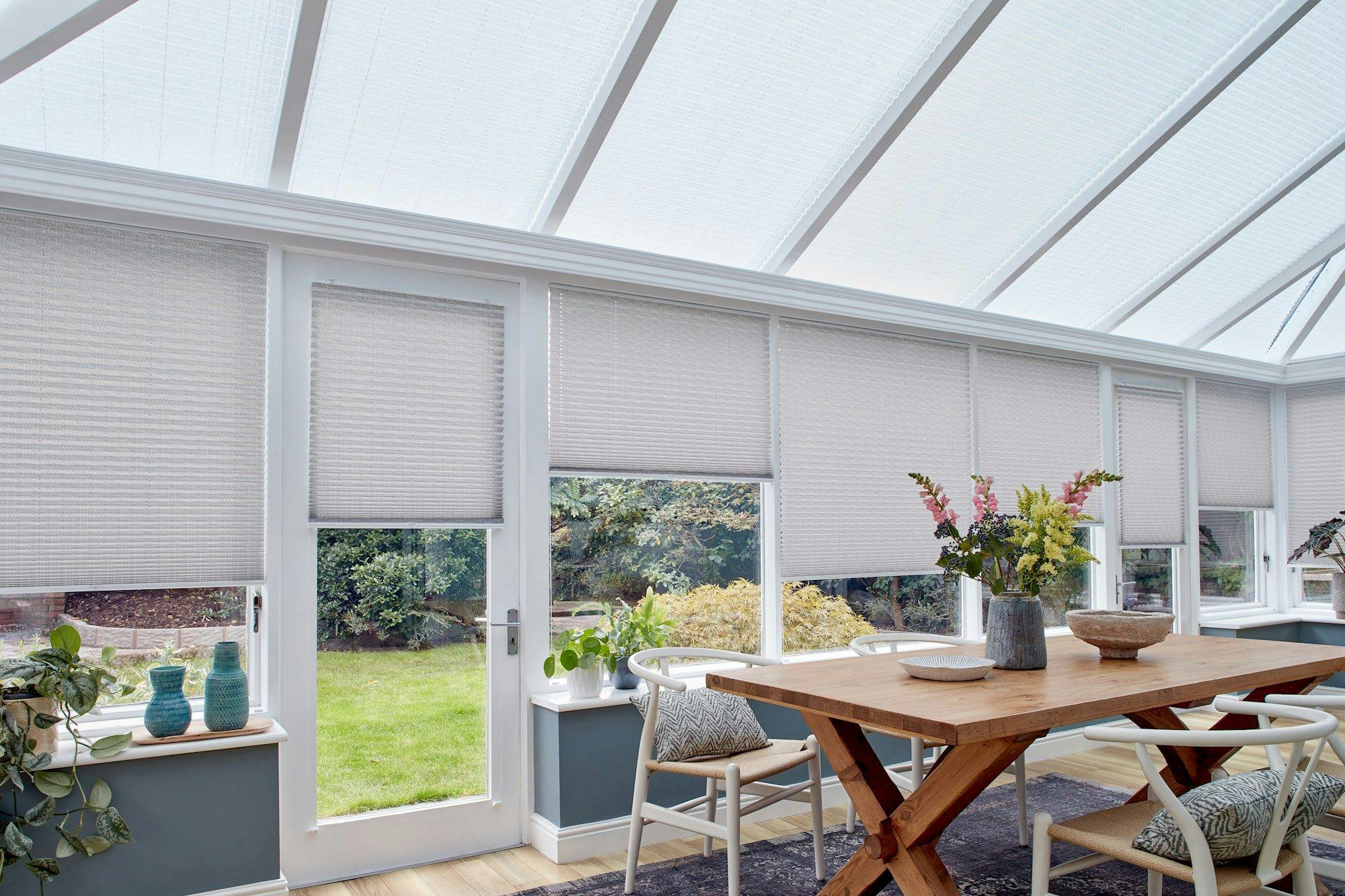 crush silver grey conservatory pleated blinds by a wooden dining table and numerous plants