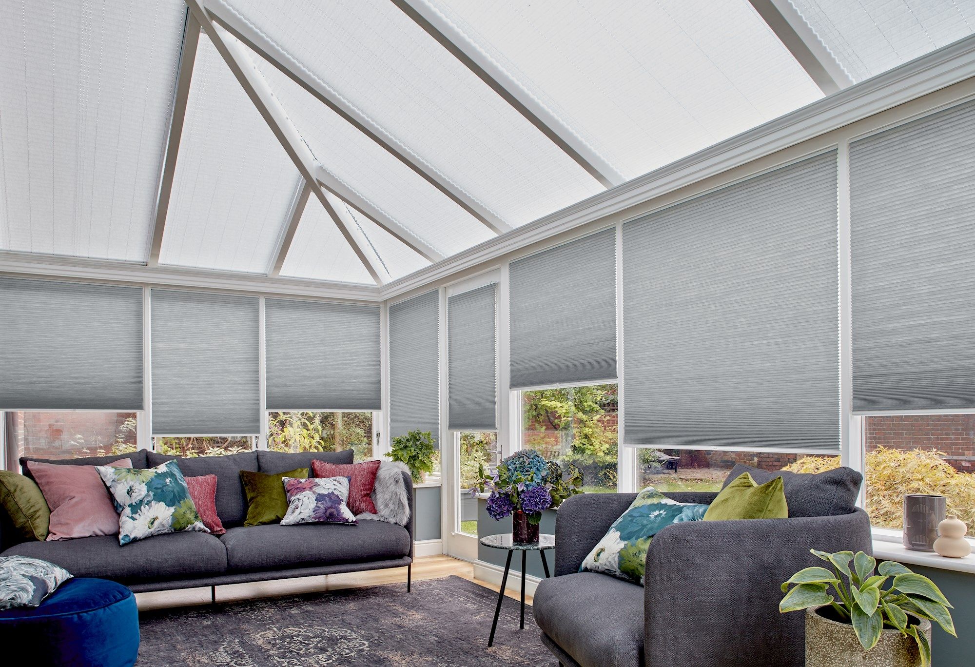 Couches adorned with colorful cushions sit under a sunlit, glass-roof conservatory. Surrounding blinds filter light, with potted plants and a small table enhancing the cozy setting.