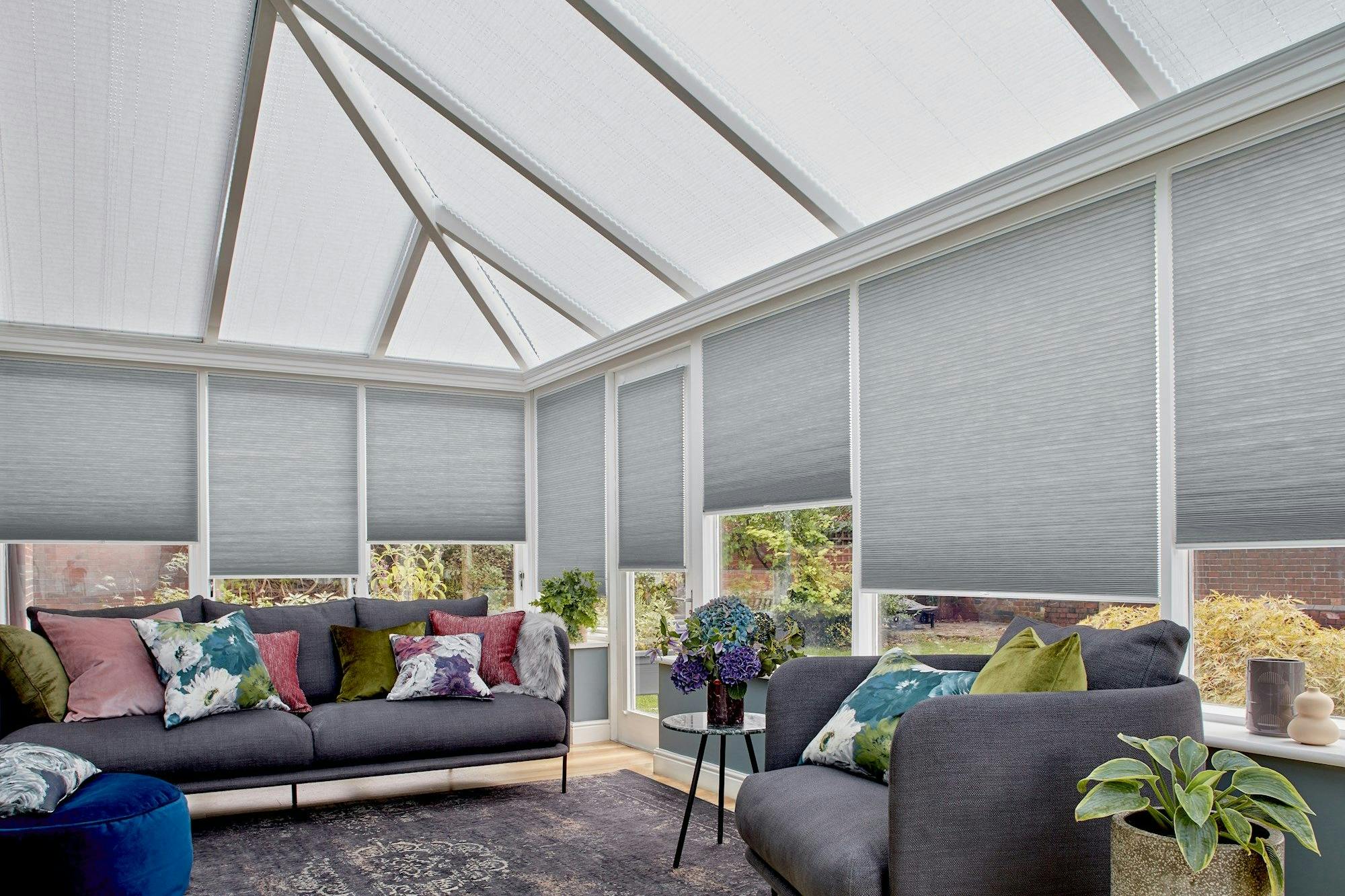 Couches adorned with colorful cushions sit under a sunlit, glass-roof conservatory. Surrounding blinds filter light, with potted plants and a small table enhancing the cozy setting.