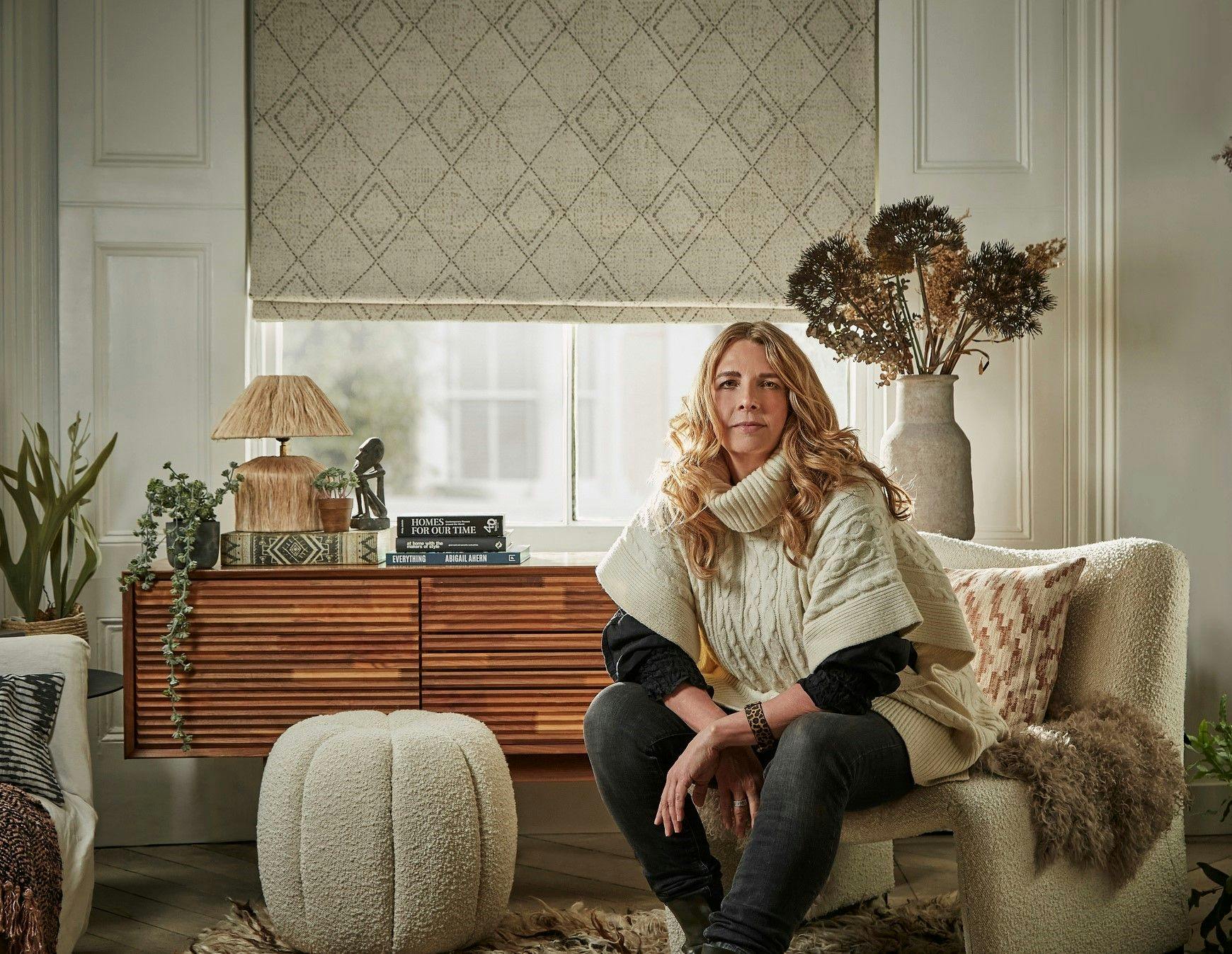 A woman with long hair sits on a cream chair, while books titled "Homes for Our Time" and "Everything" rest on a wooden cabinet adorned with plants.