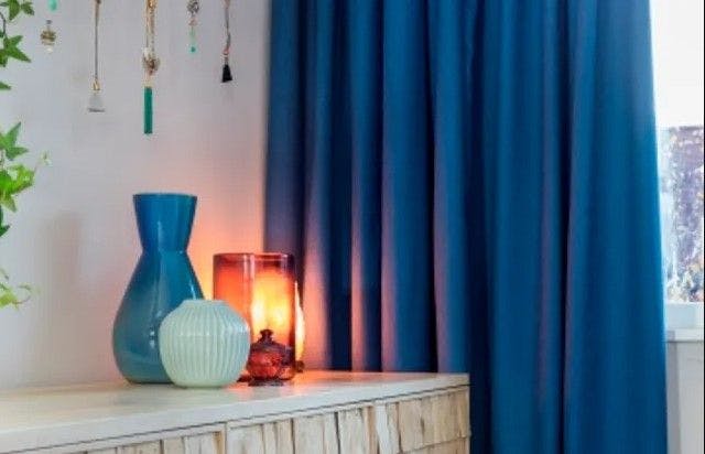 Vases rest on a wooden table next to a glowing candle, surrounded by blue curtains and hanging ornaments, creating a cozy and decorative interior scene.