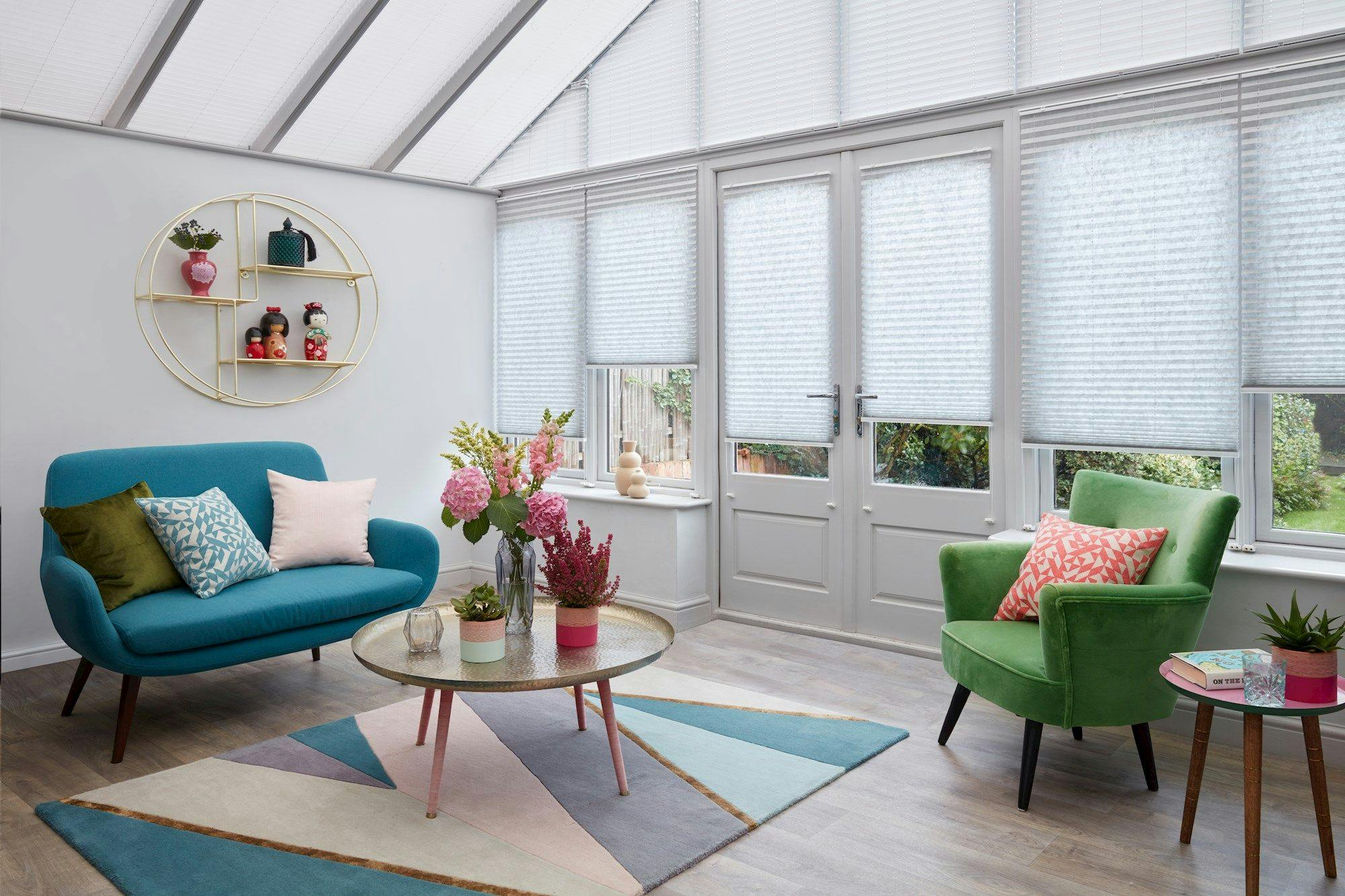 A cozy sunlit room features a teal loveseat with pillows, a circular wall shelf with decorative items, and a coffee table with flowers. Large windows, adorned with blinds, overlook greenery.