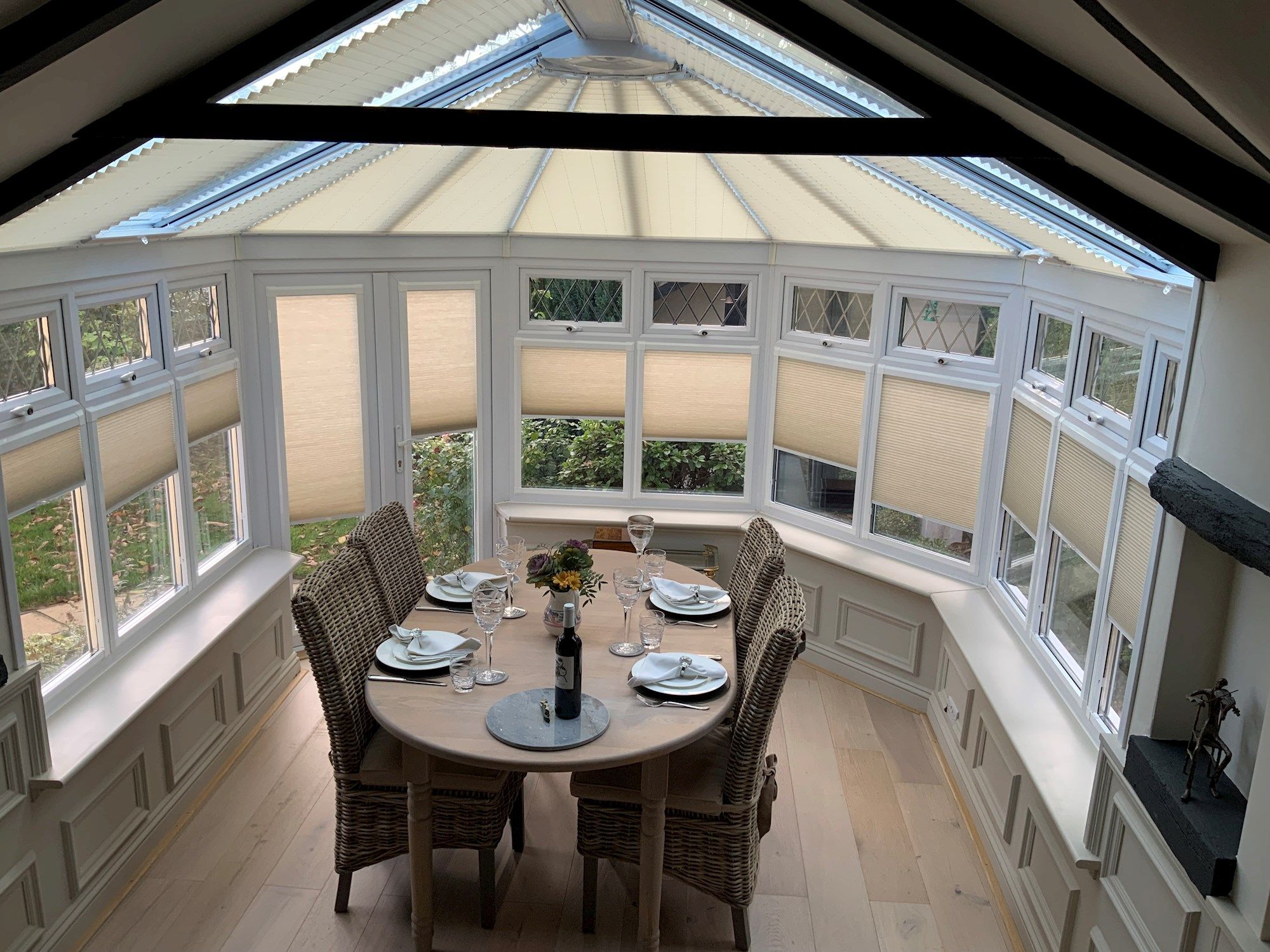 A round dining table set for four with plates, glasses, and a wine bottle, situated in a bright conservatory with wicker chairs and large windows with beige blinds.
