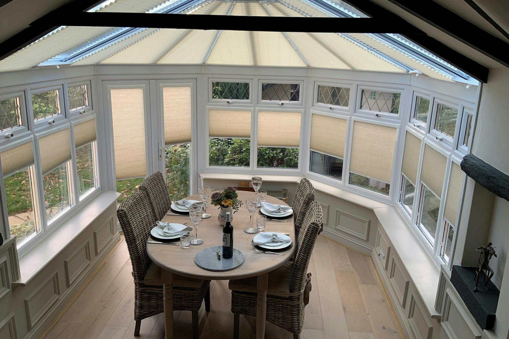 A round dining table set for four with plates, glasses, and a wine bottle, situated in a bright conservatory with wicker chairs and large windows with beige blinds.
