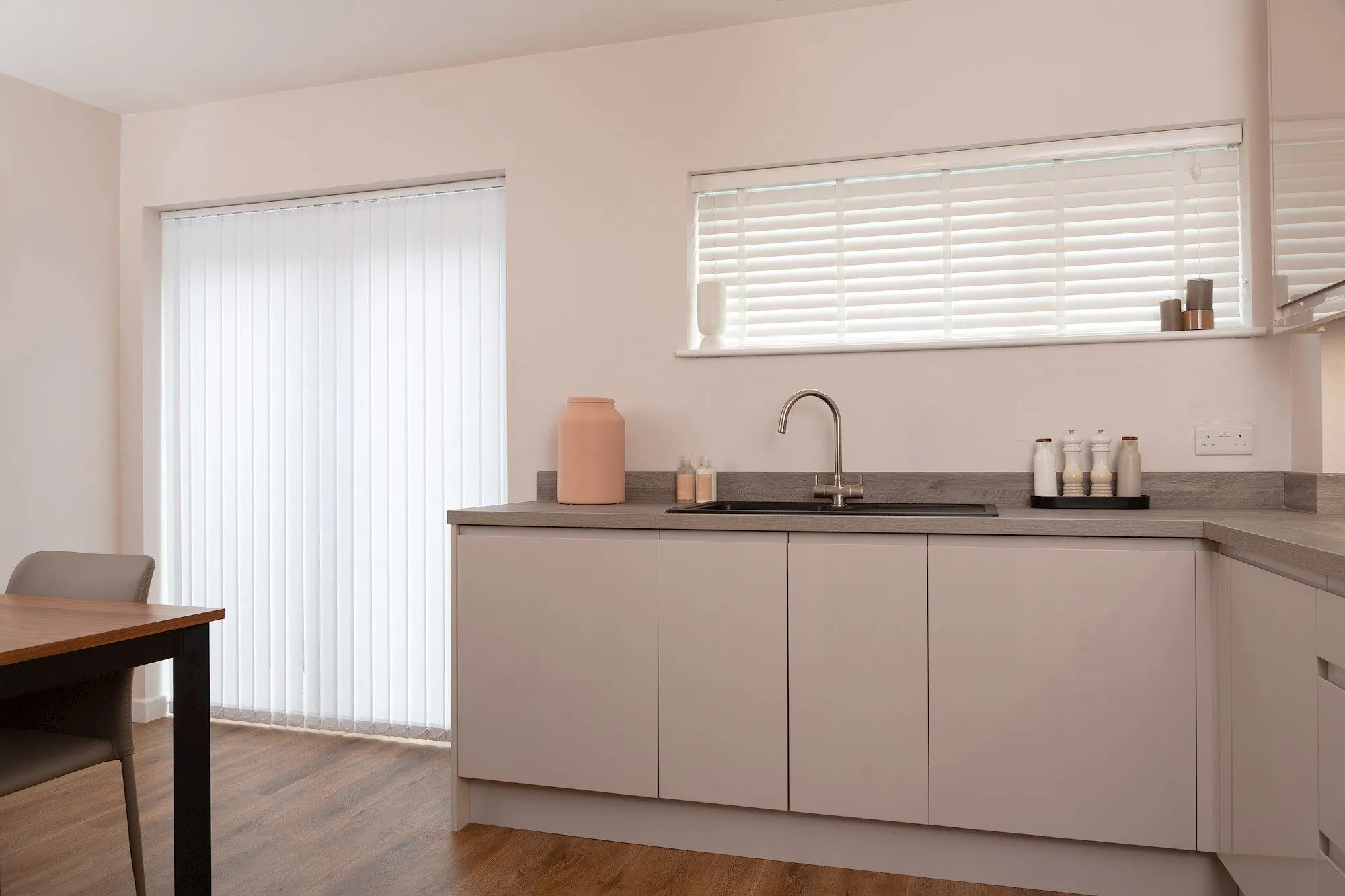 Modern kitchen with a wide patio door and window fitted with blinds, bringing in soft daylight above pale units and a sleek worktop.