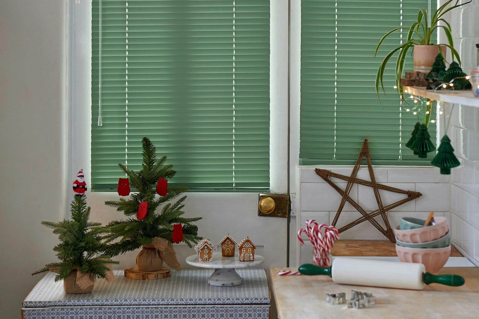 Small decorative Christmas trees adorned with red ornaments sit on a patterned surface, surrounded by gingerbread houses, candy canes, and kitchen items, set against green blinds and tiled walls.