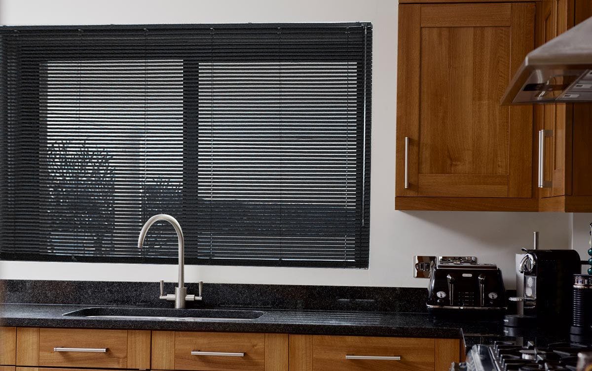A kitchen sink sits below closed black blinds, surrounded by wooden cabinets and a countertop holding a toaster and coffee machine.