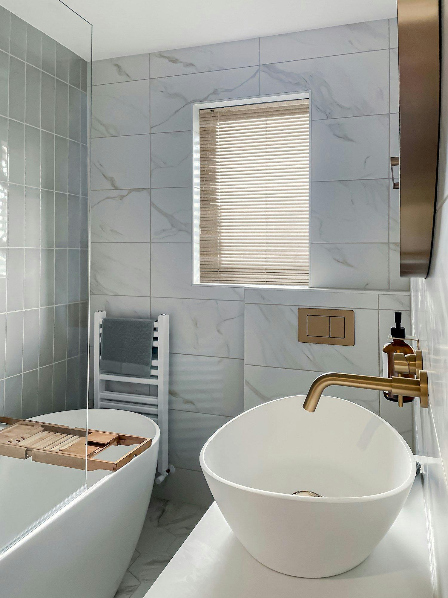 A modern bathroom features a white vessel sink with a brass faucet, situated near a bathtub equipped with a wooden tray. The space has marble-patterned tiles and a window with blinds.