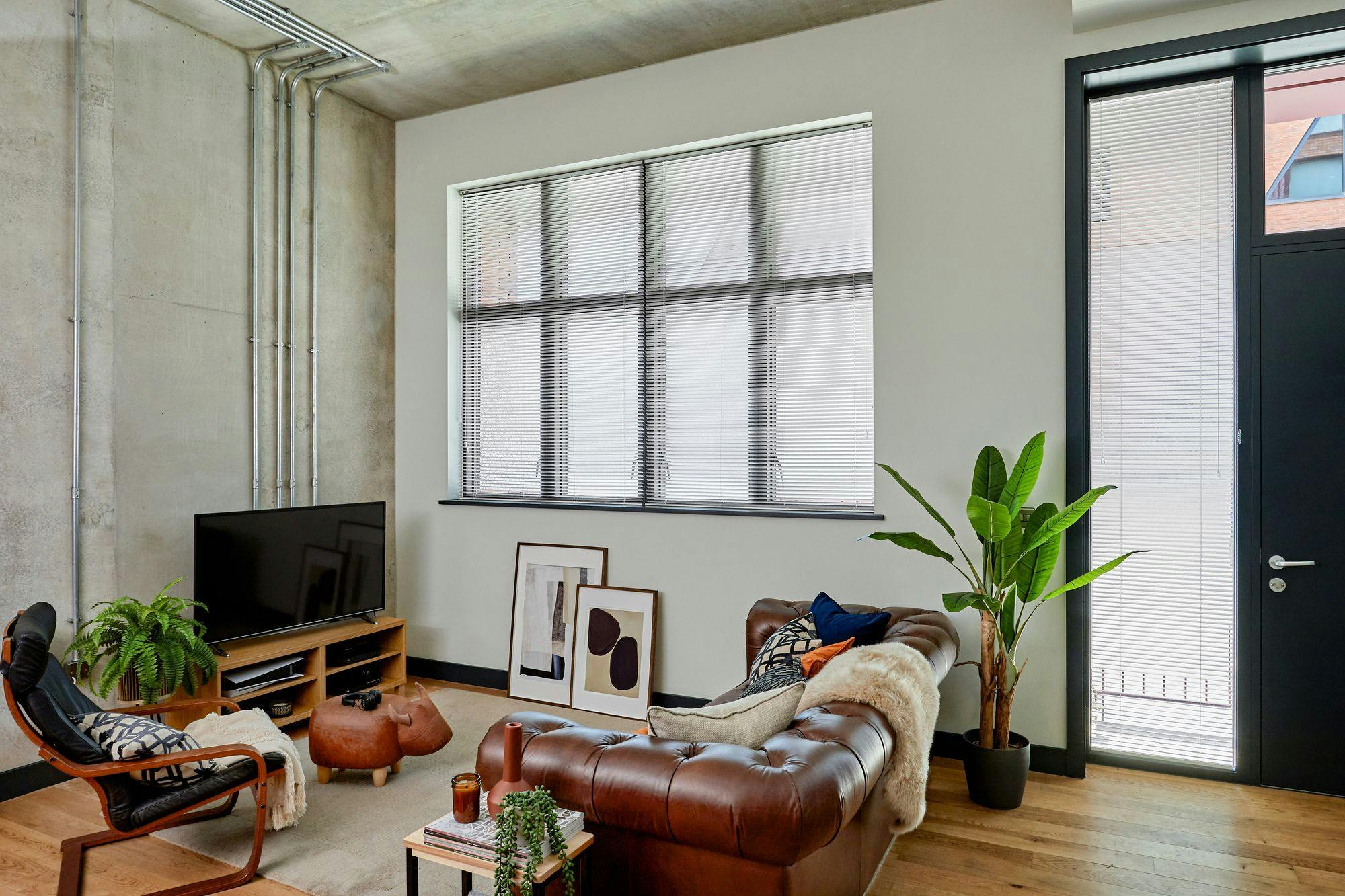 A cozy living room features a brown leather sofa adorned with cushions, a modern chair, and a wooden TV stand. Large windows with blinds and plants enhance the airy space.