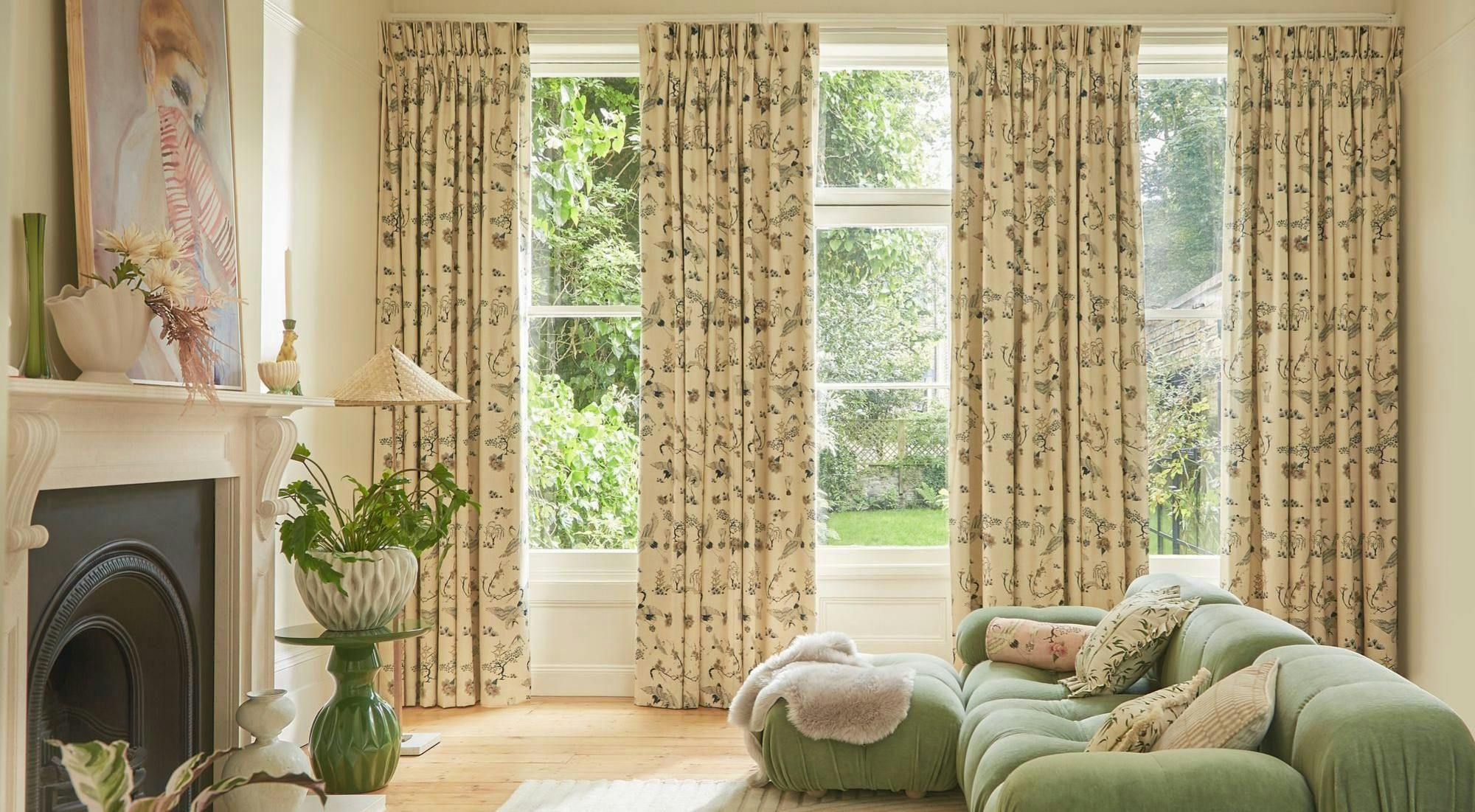 A green sofa sits in a living room with patterned curtains and large windows showing a garden. A fireplace with vases and an artwork decorates the space, adding warmth and style.
