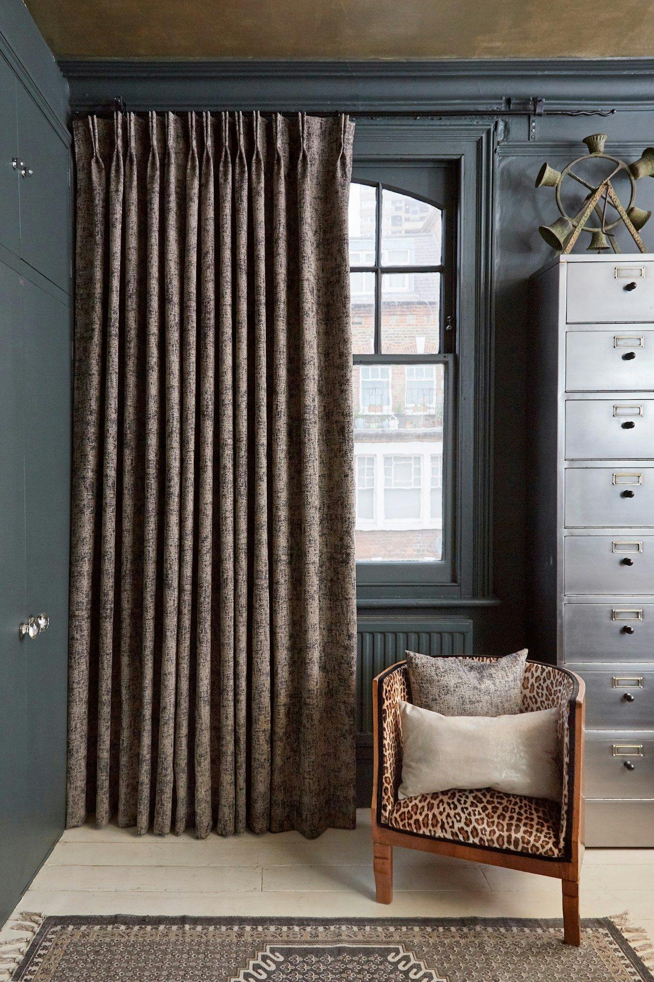 Curtains hang beside a window, partially open, in a room with dark green walls. A leopard-print chair with cushions sits on a patterned rug near a tall, gray metal filing cabinet.