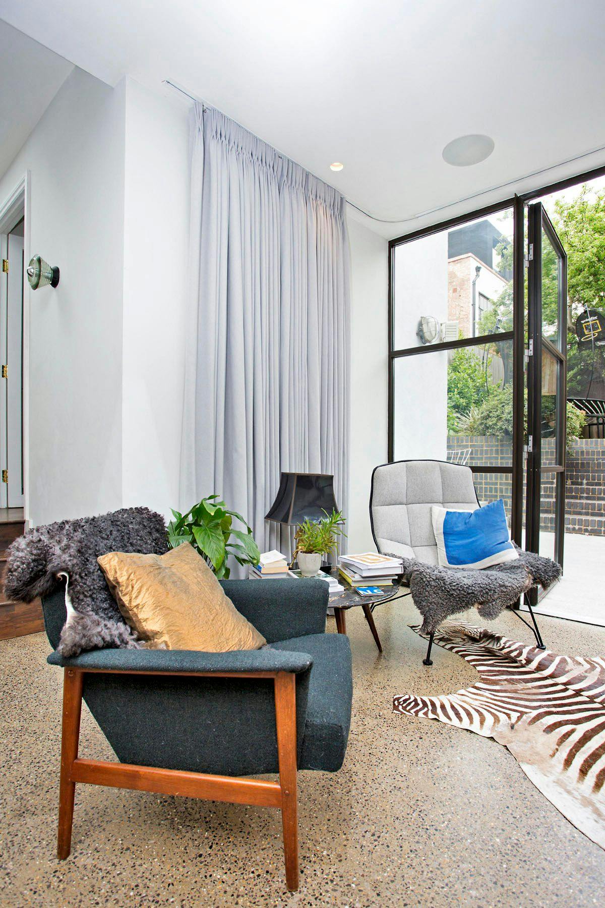 Armchairs adorned with cushions and throws sit on a zebra-print rug in a bright room with large windows and a gray curtain. Nearby, a small table holds books and a plant.