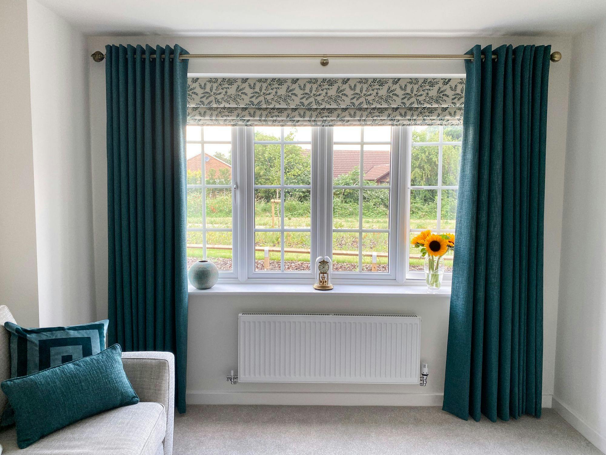 Window displaying teal curtains, partially open; vase with sunflowers and a decorative globe on the sill. Couch with teal cushions beside, overlooking a green garden.