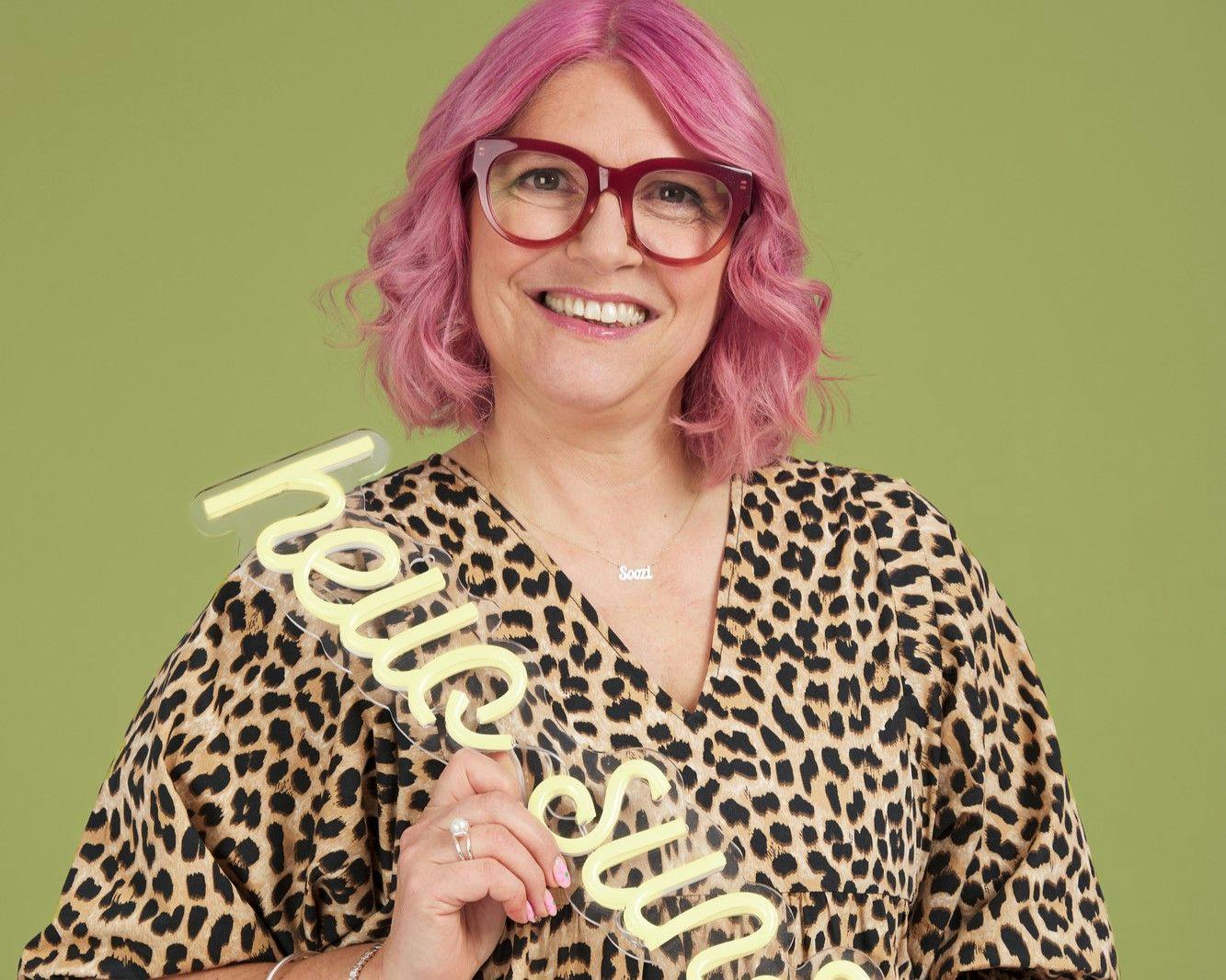 A person with pink hair holds a sign reading "hello star," wearing glasses and a leopard-print outfit, against a green background. A necklace with the name "Soozi" is visible.