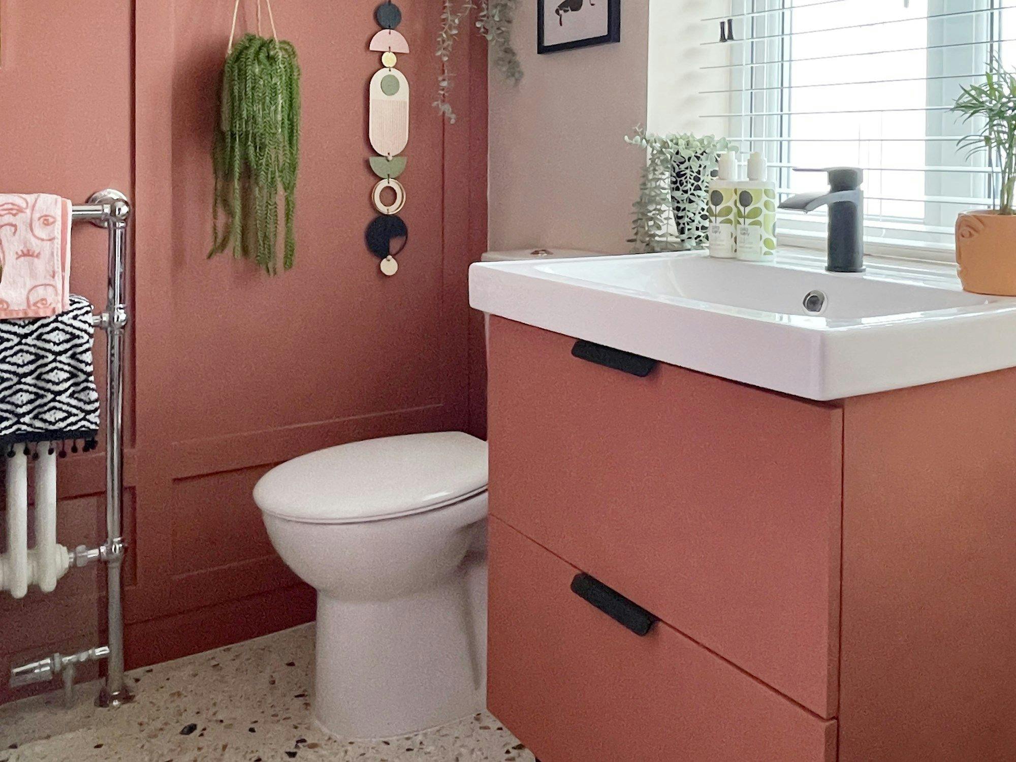 A white toilet beside a modern rust-colored vanity holds a sink with a black faucet. Hanging plants and geometric wall art decorate the bathroom with patterned towels on a steel rack.