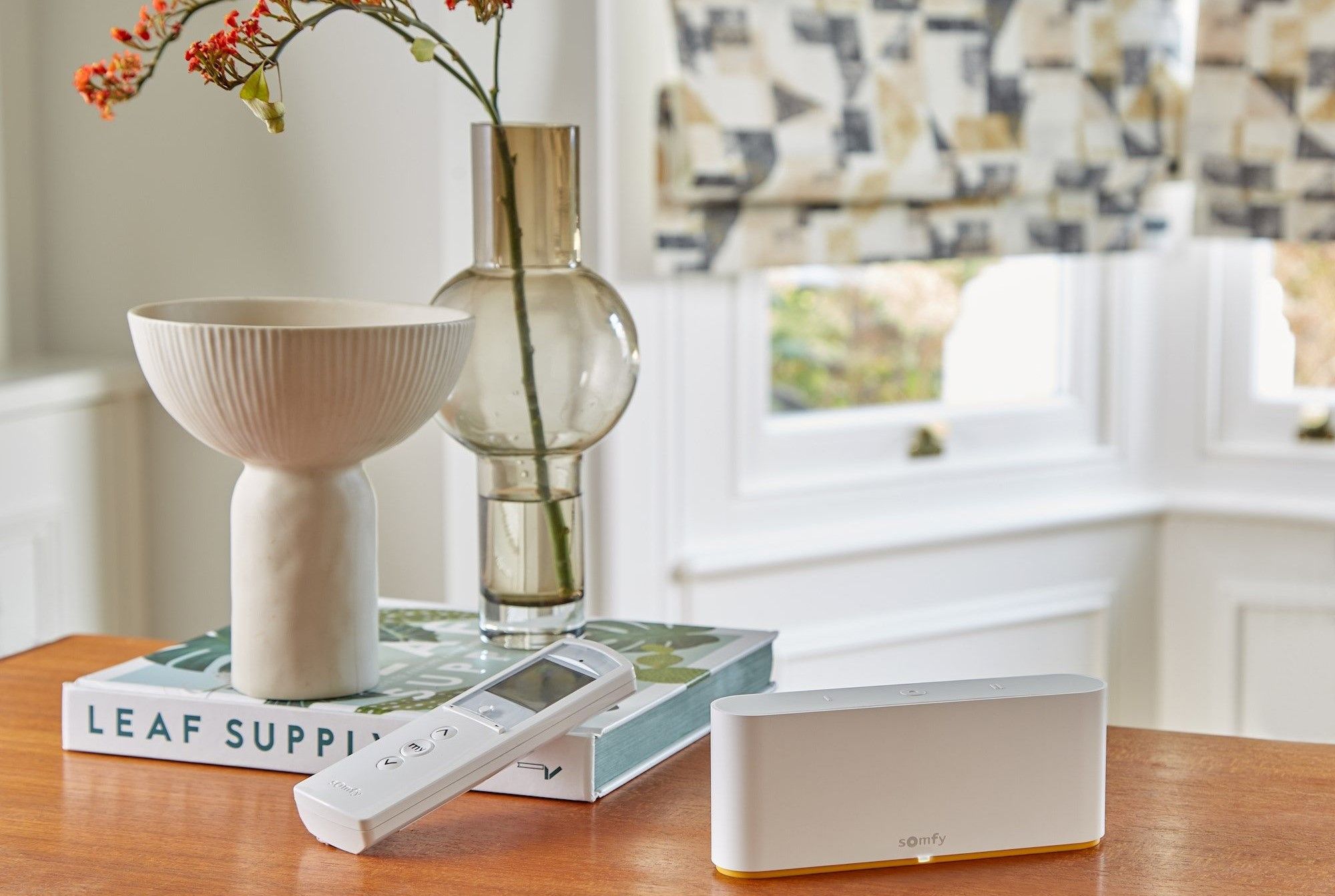 A white remote and Somfy device rest on a wooden table in a room. Nearby is a ceramic bowl on books labeled "LEAF SUPPLY" and a glass vase with flowers by a window.