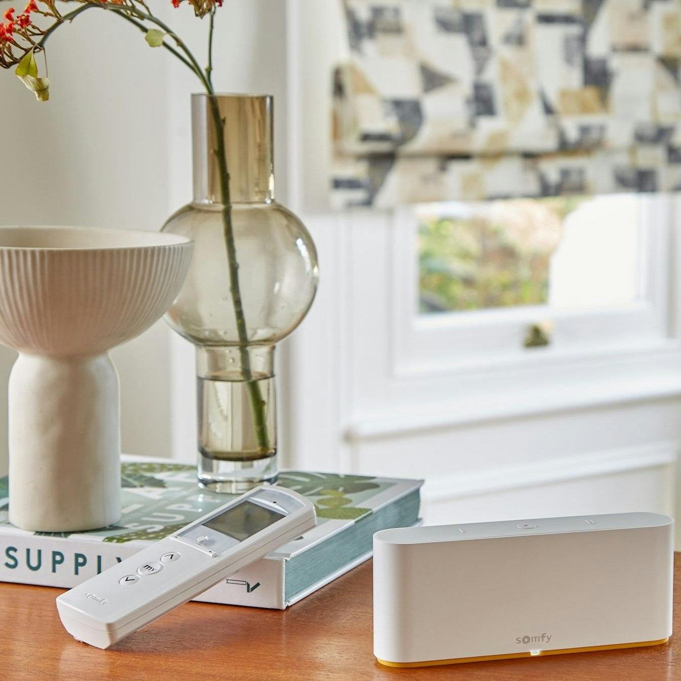 A white remote and Somfy device rest on a wooden table in a room. Nearby is a ceramic bowl on books labeled "LEAF SUPPLY" and a glass vase with flowers by a window.
