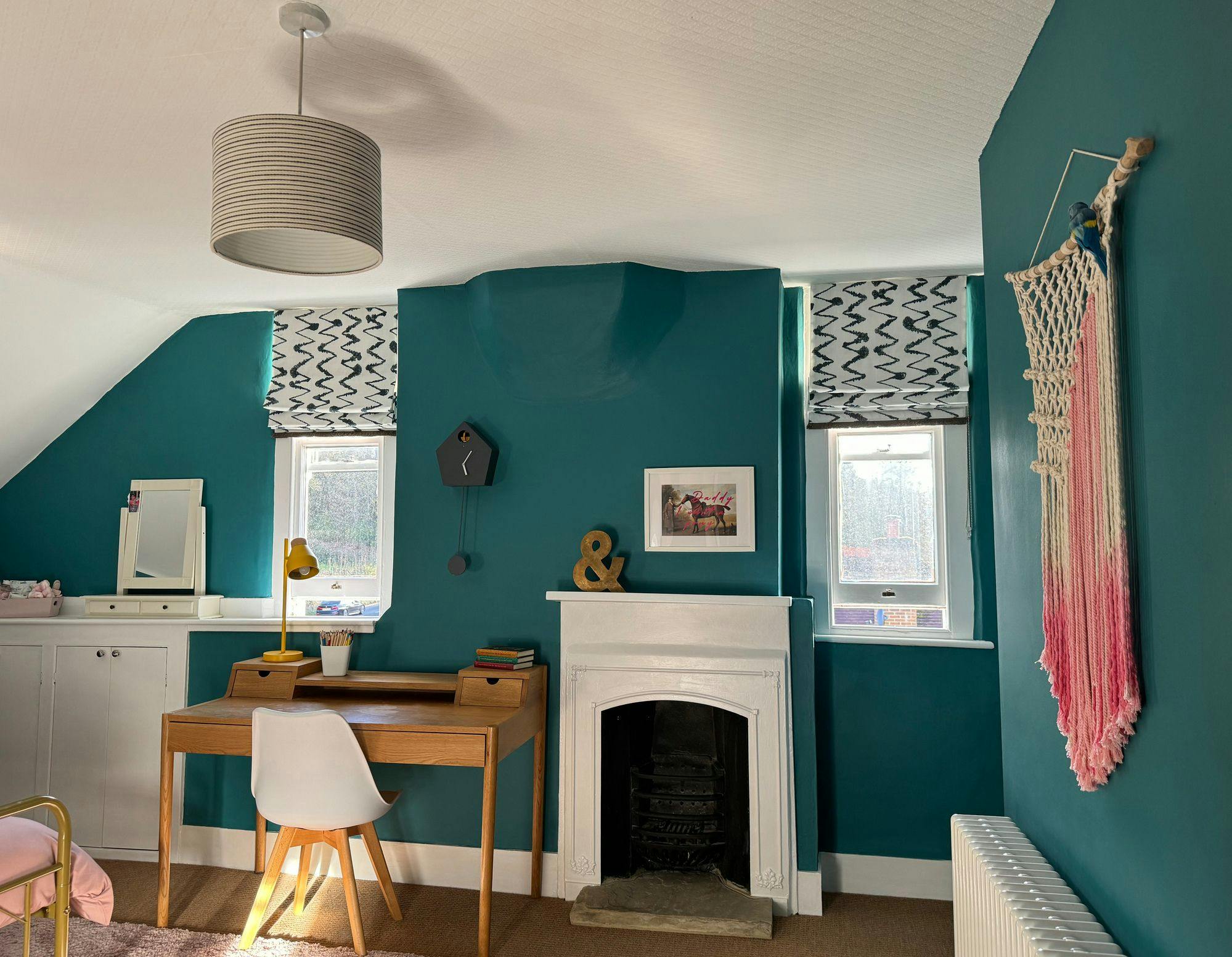 Desk with chair and lamp sits against a teal wall between windows, next to a white fireplace. Decor includes a wall hanging and a picture frame with horses.