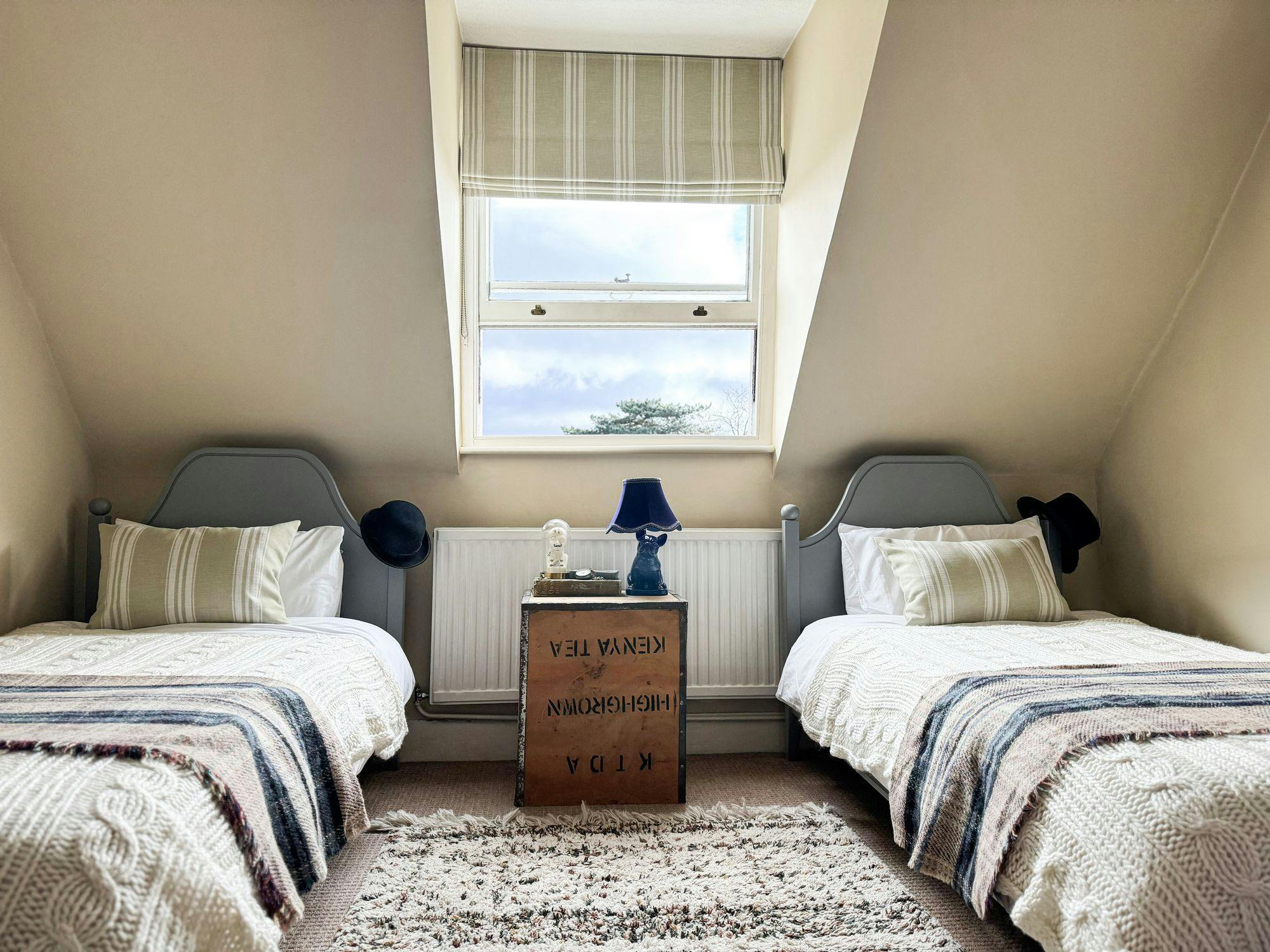 Two single beds with knitted blankets and striped pillows sit on either side of a box labeled "KENYA TEA" under a window in a cozy, attic-style room.