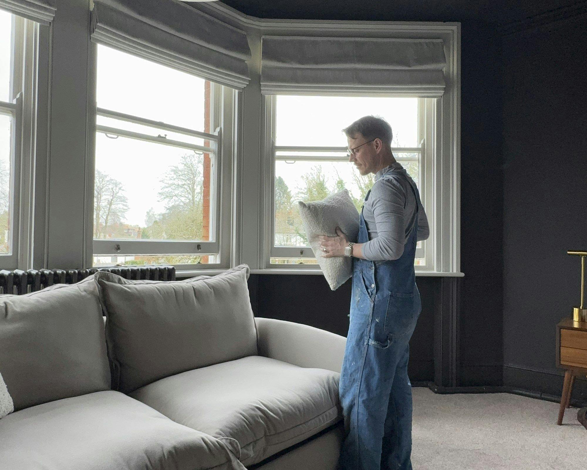 A person wearing overalls arranges a cushion on a sofa. They are in a living room with large bay windows, letting in natural light and revealing trees outside.