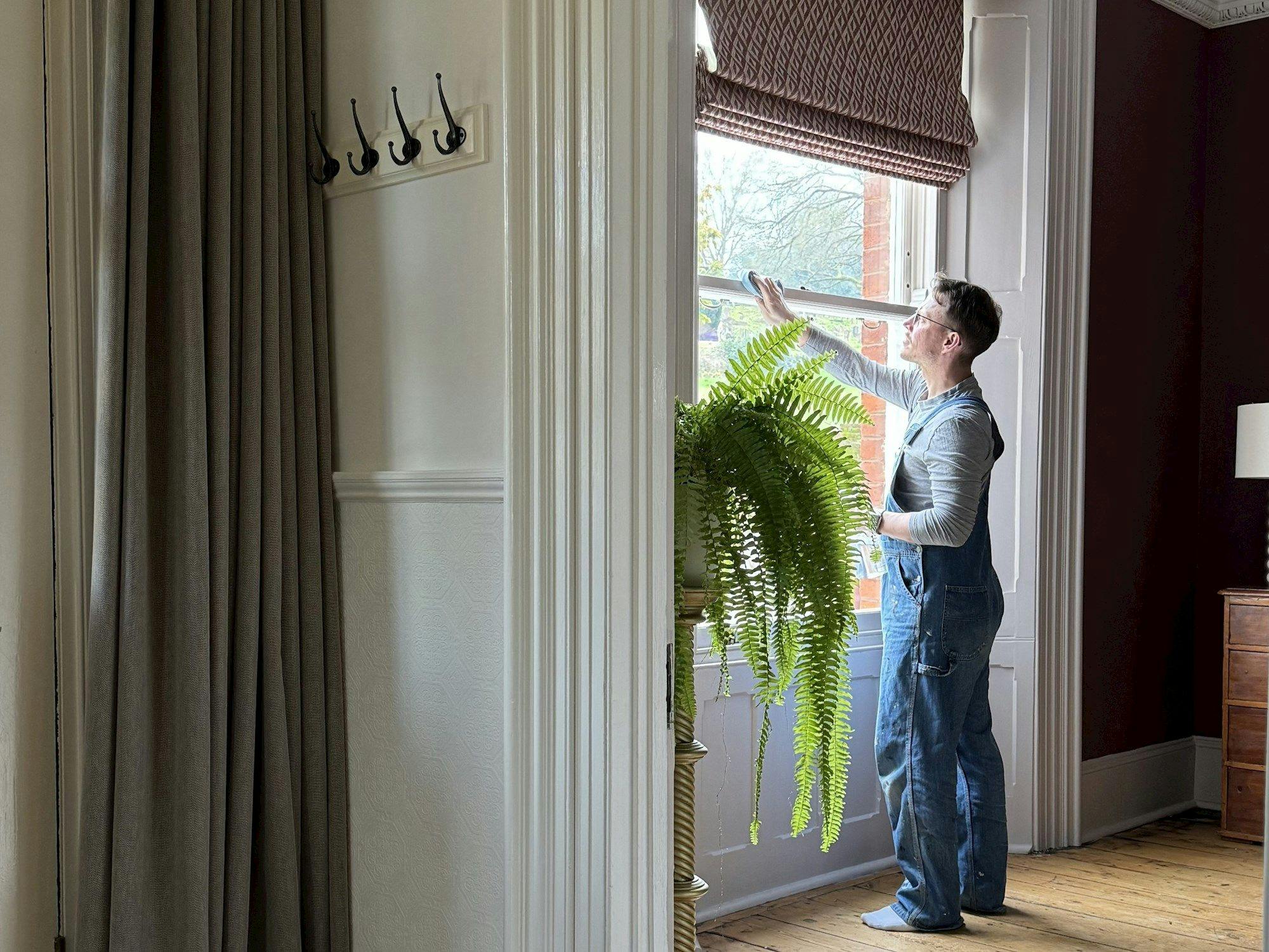 A person in overalls cleans a tall window, light streaming in, next to a lush fern in a spacious room with wooden flooring and a visible coat rack.