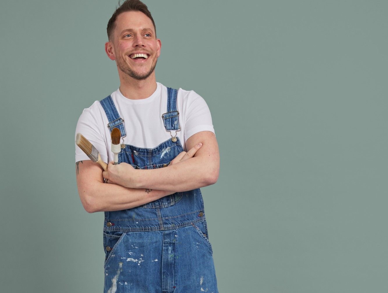 A person in blue paint-splattered overalls, smiling and crossing their arms, holds paintbrushes against a plain green background.