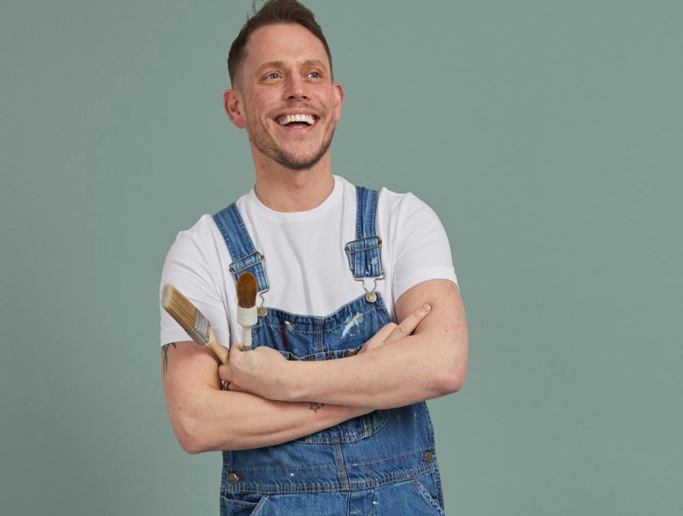 A person in blue paint-splattered overalls, smiling and crossing their arms, holds paintbrushes against a plain green background.