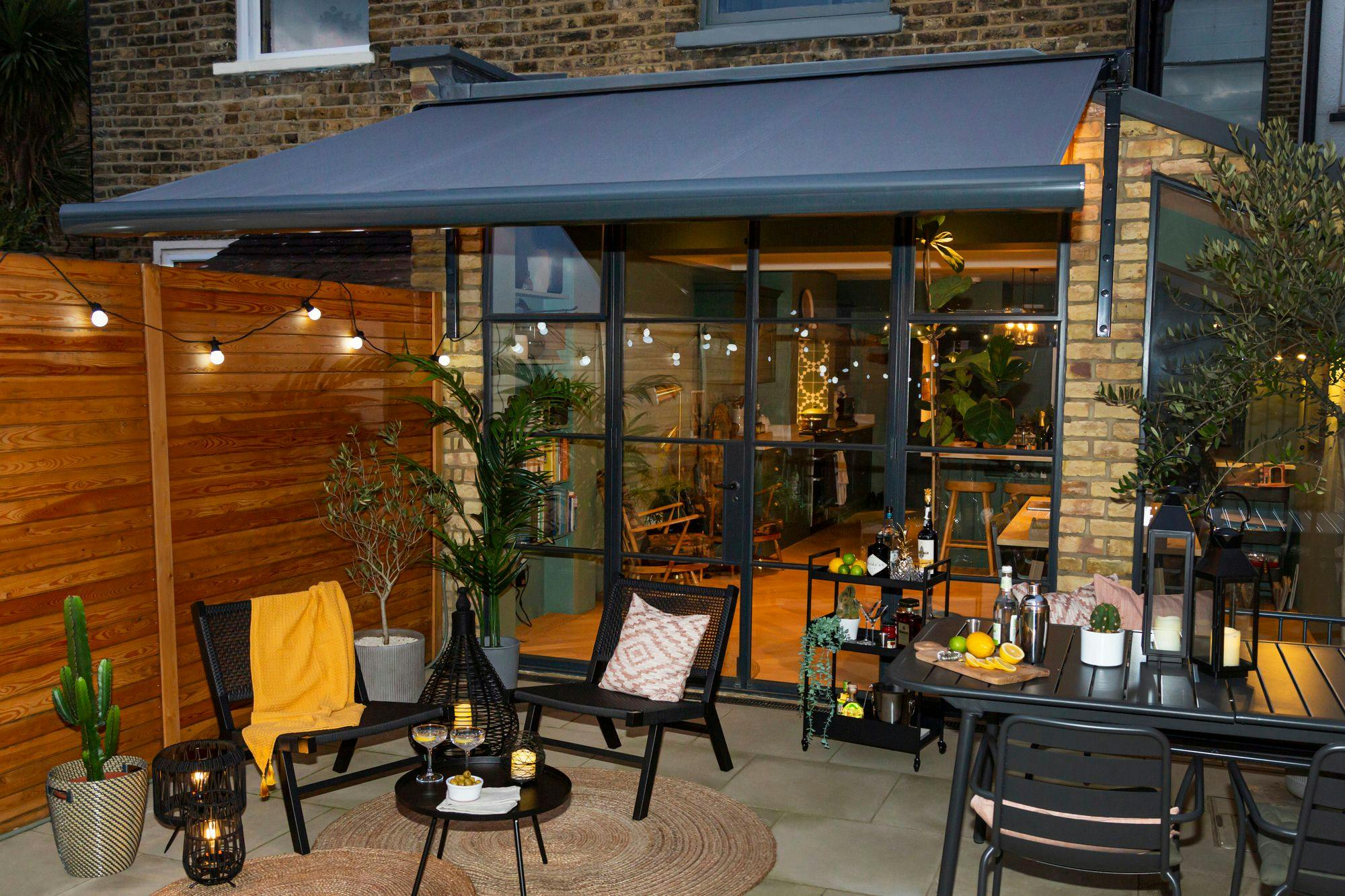 Patio chairs are arranged with a table holding drinks and snacks. A wooden fence with string lights encloses the area, adjacent to a house with large glass doors revealing an illuminated interior.