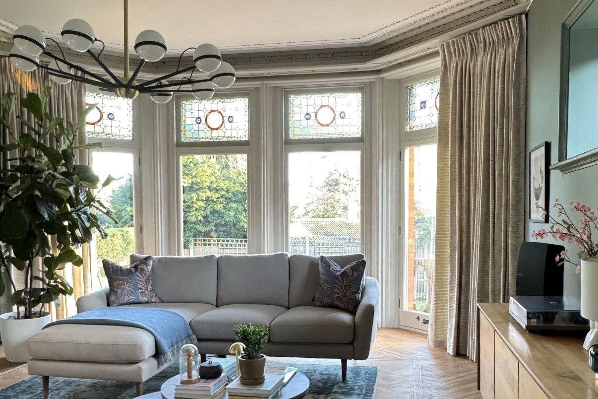 A modern living room features a grey sectional sofa with decorative pillows and a blue throw, positioned in front of large windows with stained glass. A chandelier hangs above a coffee table with books and plants, adjacent to a wooden sideboard with decor.