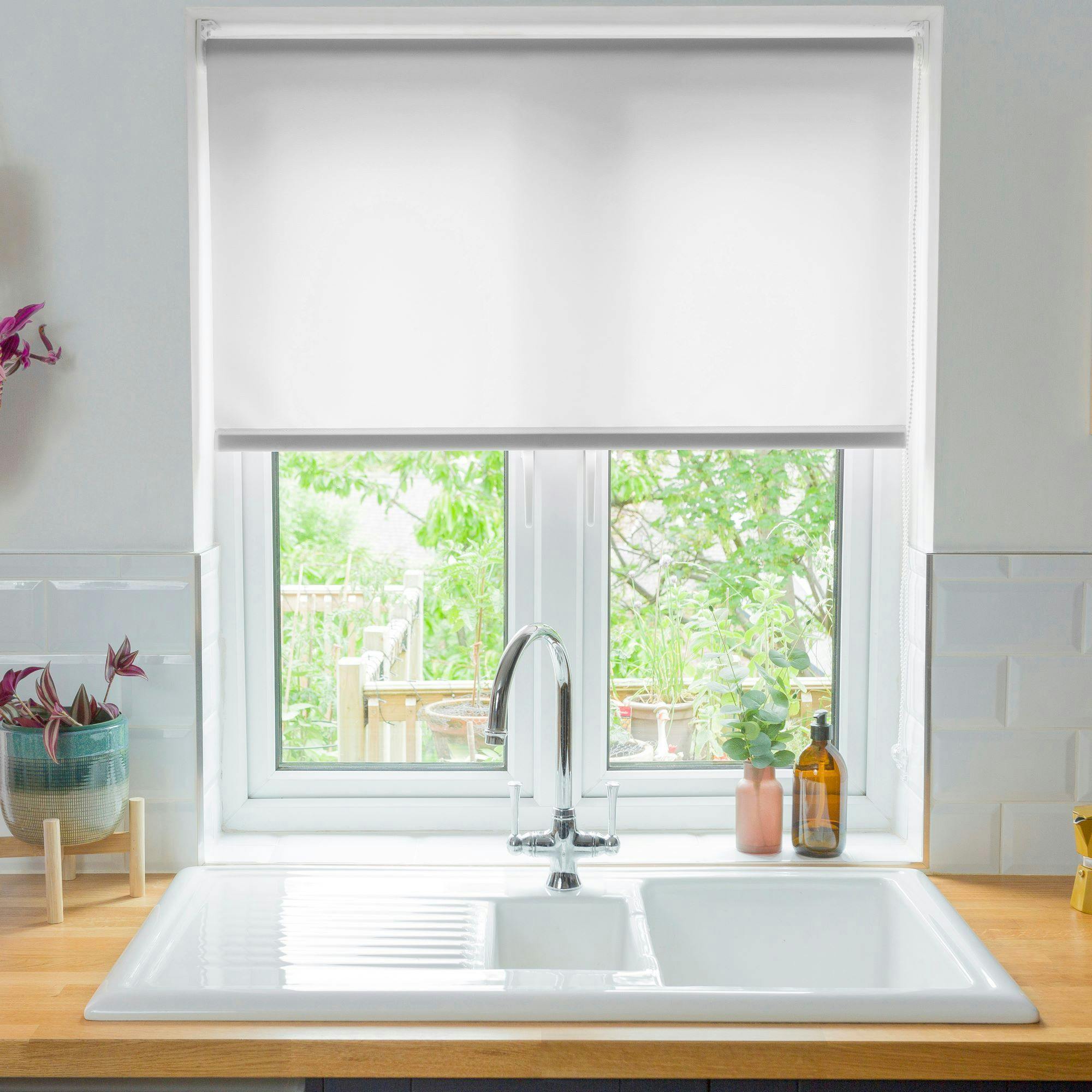 White ceramic kitchen sink with a chrome faucet sits beneath a closed window blind, surrounded by potted plants and bottles, with an outdoor garden visible through the window.