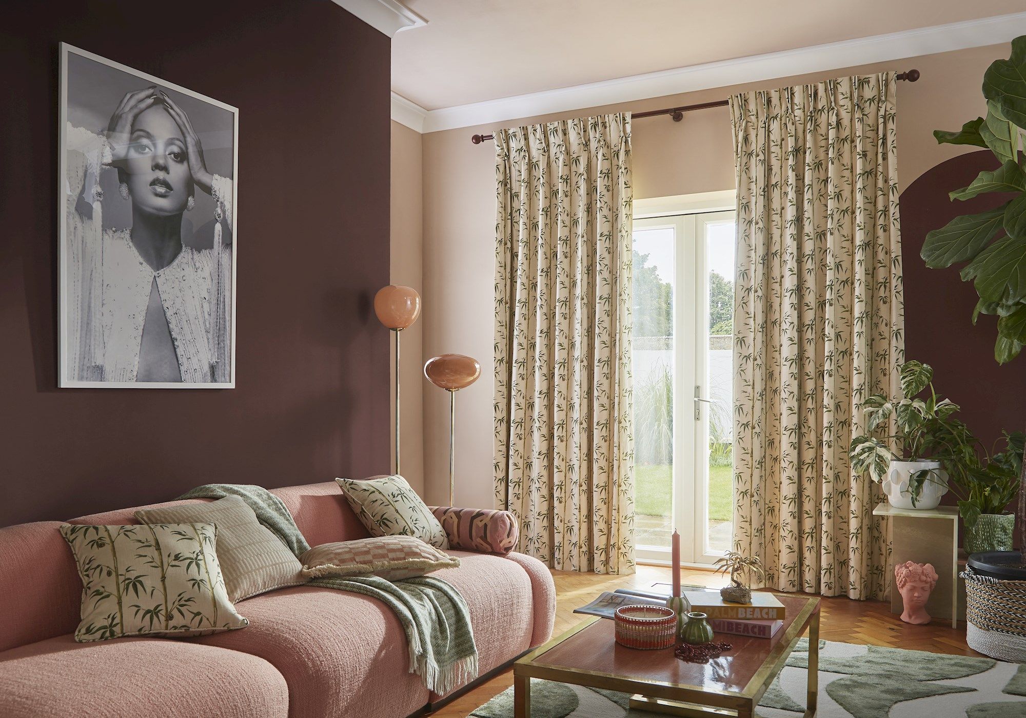 A cozy living room features a pink sofa with patterned cushions and a blanket. A large monochrome portrait hangs on a dark wall. Nearby, floor-length patterned curtains frame a glass door with a garden view. A wooden coffee table displays books and decor. Houseplants add greenery.