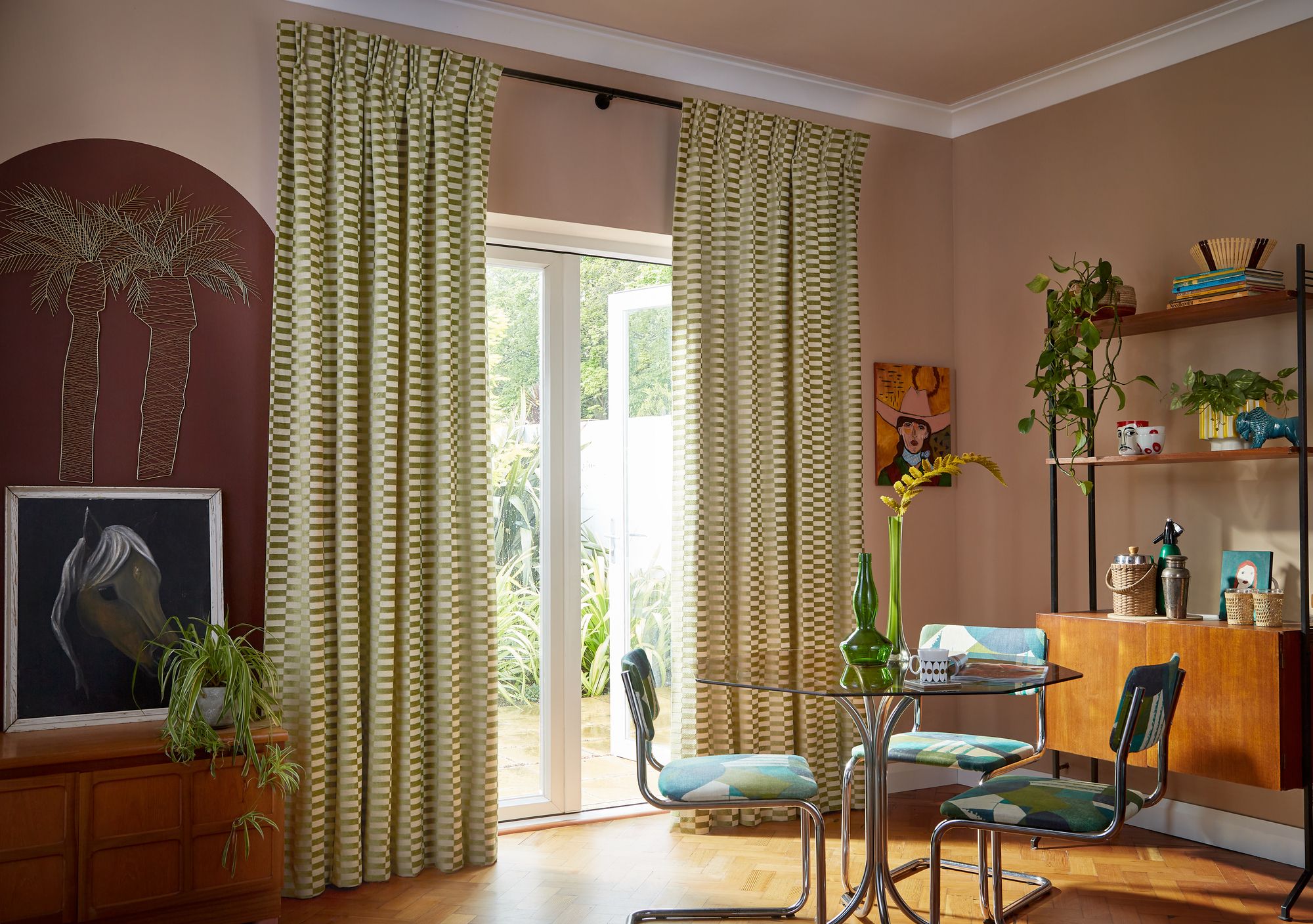 Curtains hang in front of glass doors, leading to a garden. A round table with chairs is nearby, and the room features plants, artwork, and wooden furniture.