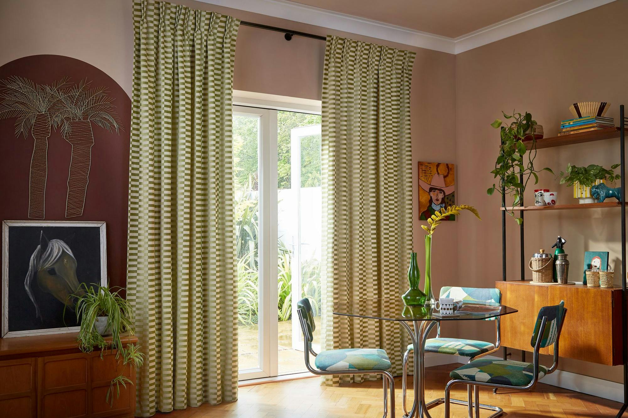 Curtains hang in front of glass doors, leading to a garden. A round table with chairs is nearby, and the room features plants, artwork, and wooden furniture.
