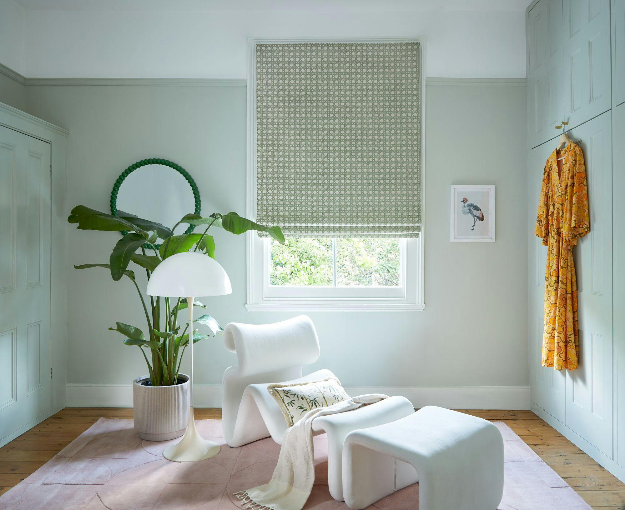 Modern white chair and footrest sit on pink rug, next to a large potted plant and white lamp. Yellow robe hangs nearby; patterned blind covers window. Bird artwork decorates mint green walls.