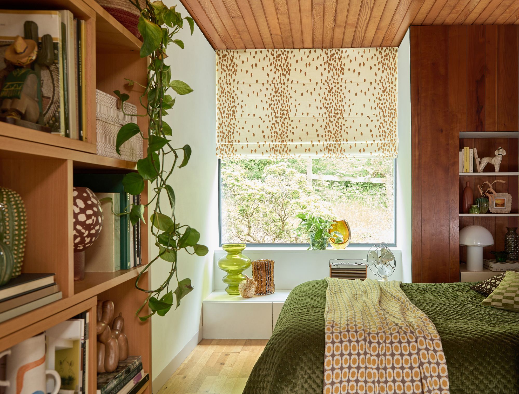 Bookshelves filled with books and decor flank a cozy bedroom. A green bedspread covers the bed, near a window with patterned blinds overlooking a garden. A plant hangs from the shelf.