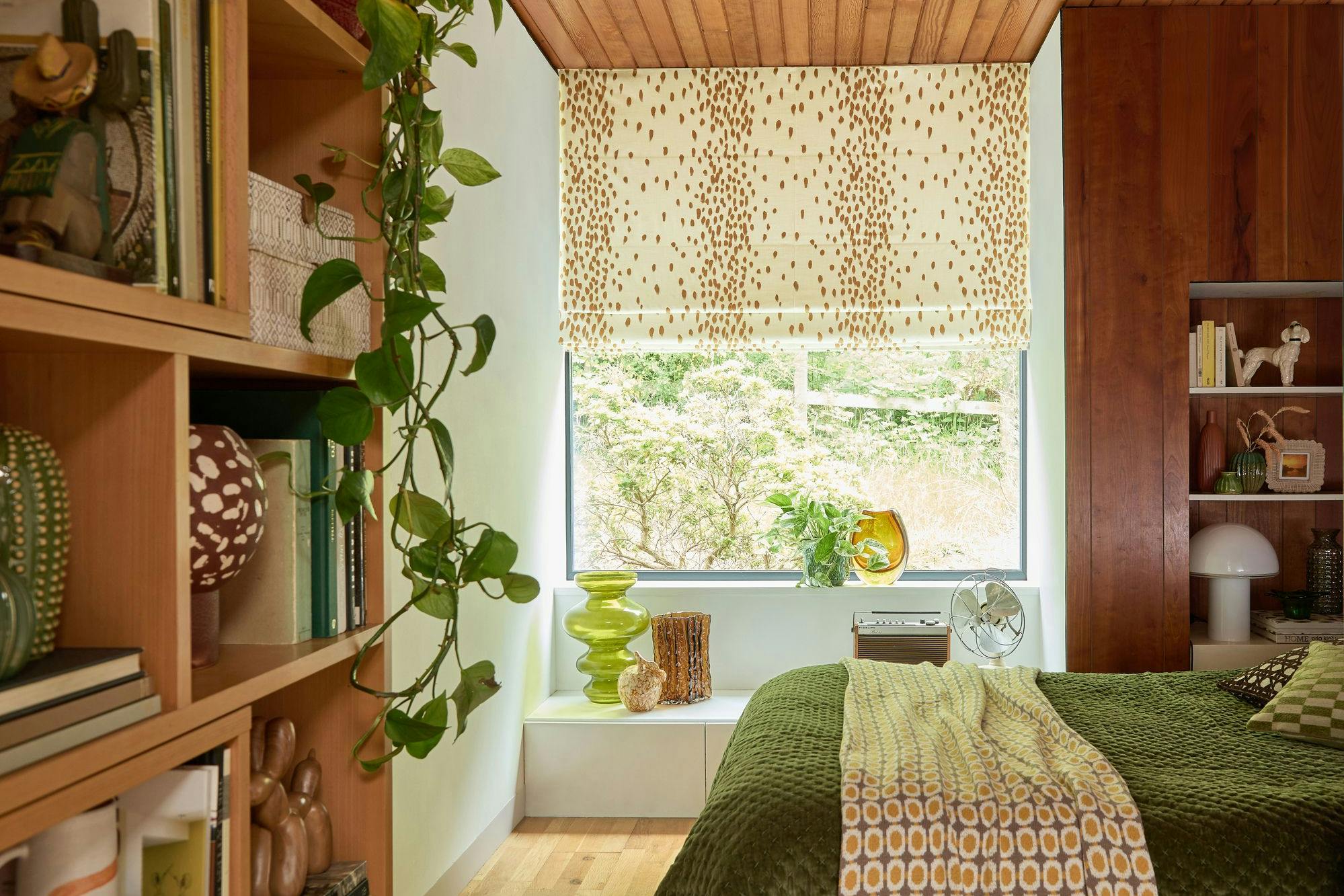Bookshelves filled with books and decor flank a cozy bedroom. A green bedspread covers the bed, near a window with patterned blinds overlooking a garden. A plant hangs from the shelf.