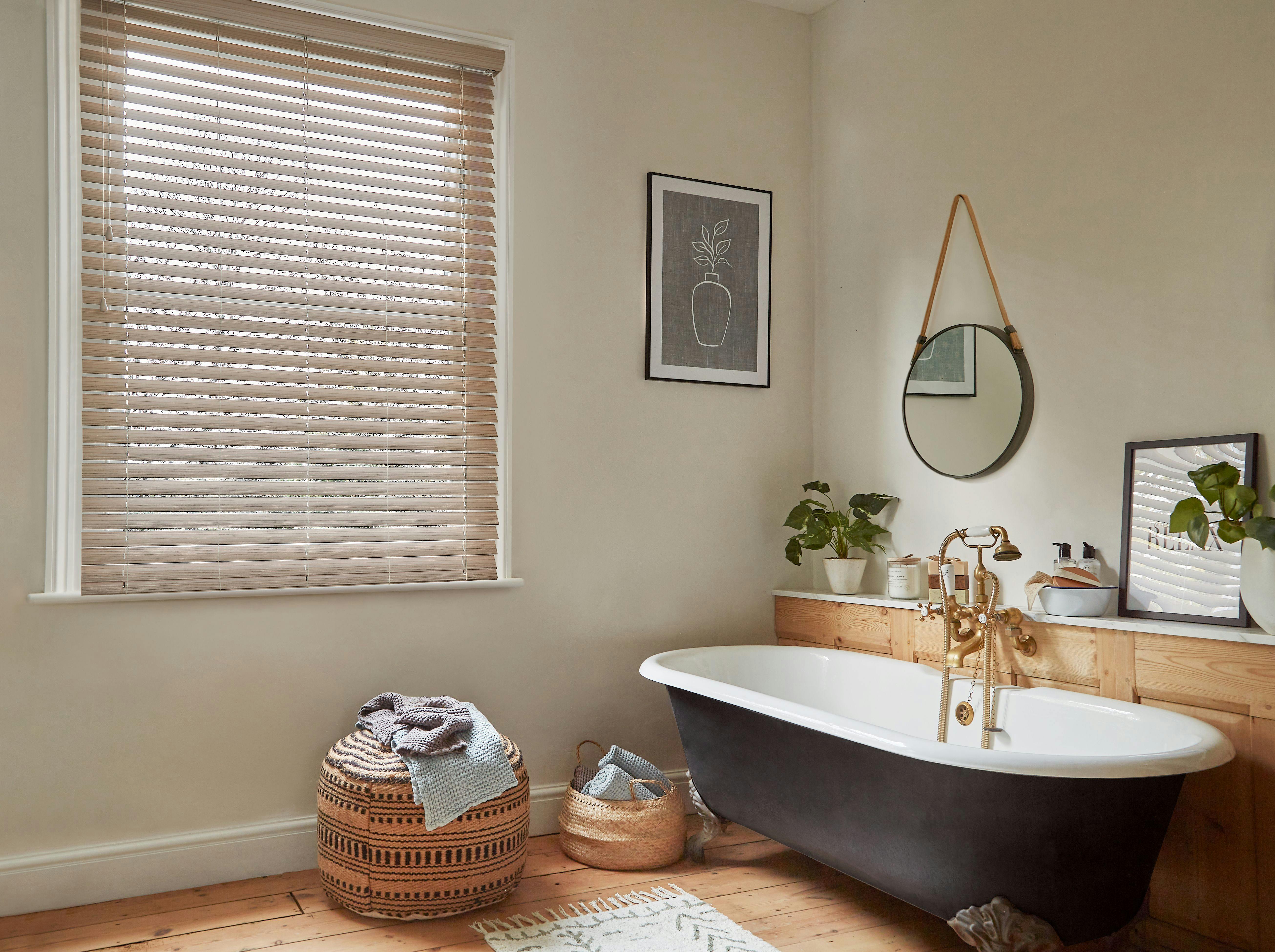 A black bathtub with gold fixtures sits in a cozy bathroom, surrounded by plants and woven baskets, against light walls with framed art and a round mirror.