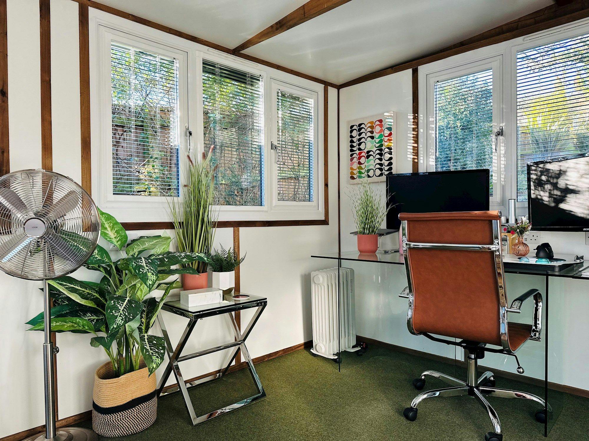 A home office features a brown leather chair at a glass desk with dual monitors. Green plants decorate the room, and sunlight filters through blinds covering large windows.